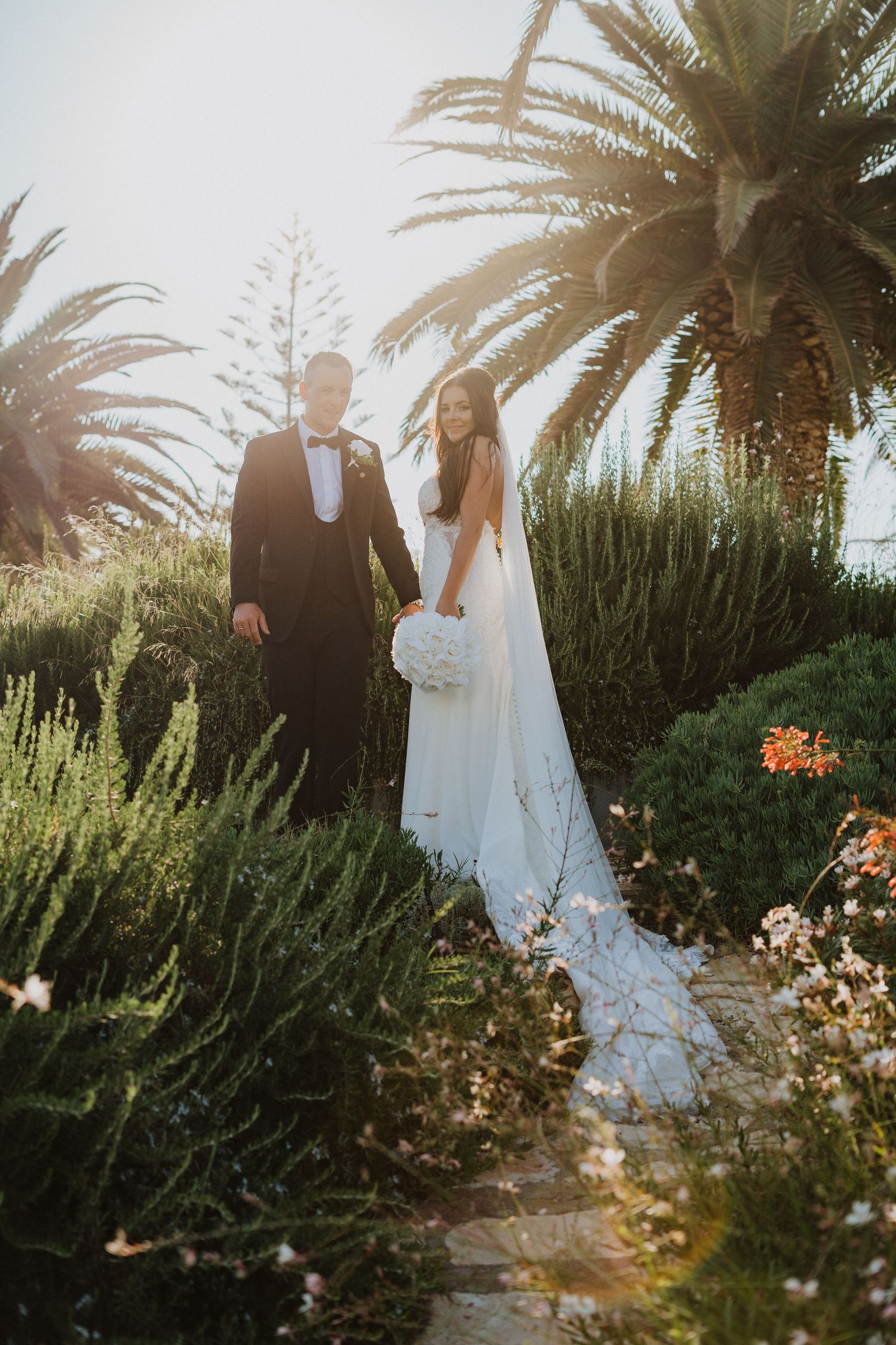A wedding couple poses in a tropical garden setting with palm trees and flowering plants.