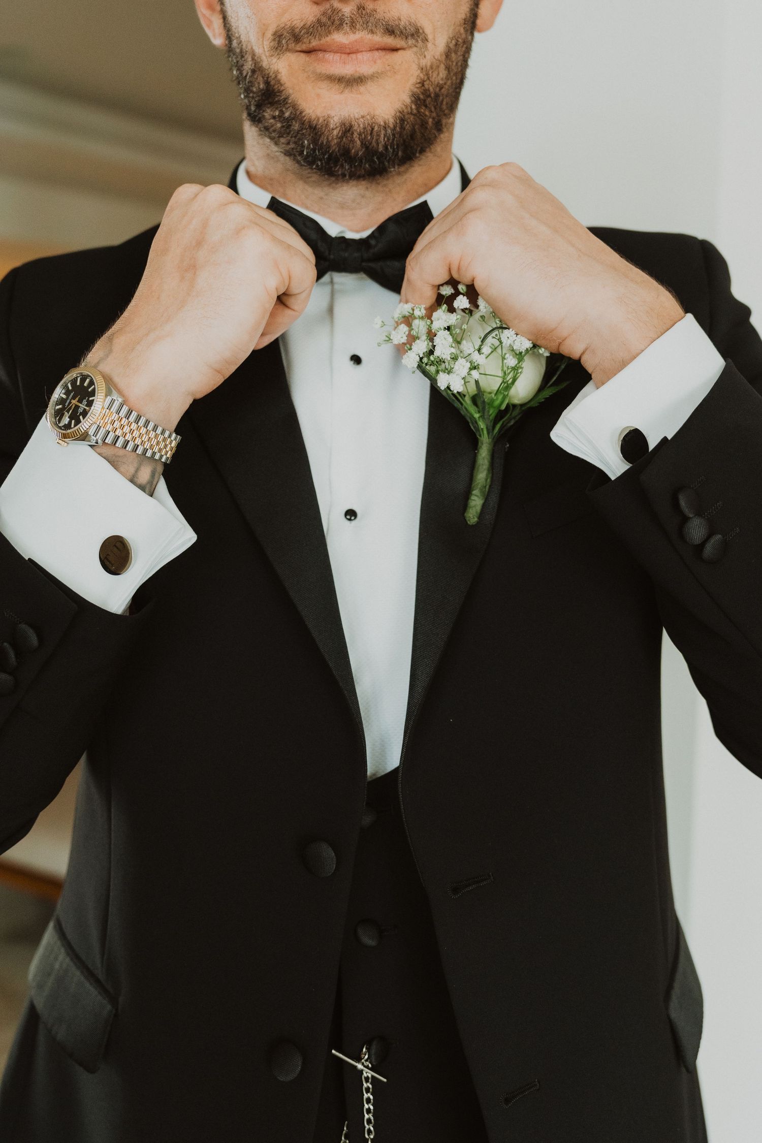 Close-up detail shots of a groom adjusting his black tuxedo bow tie and cufflinks with white boutonniere.
