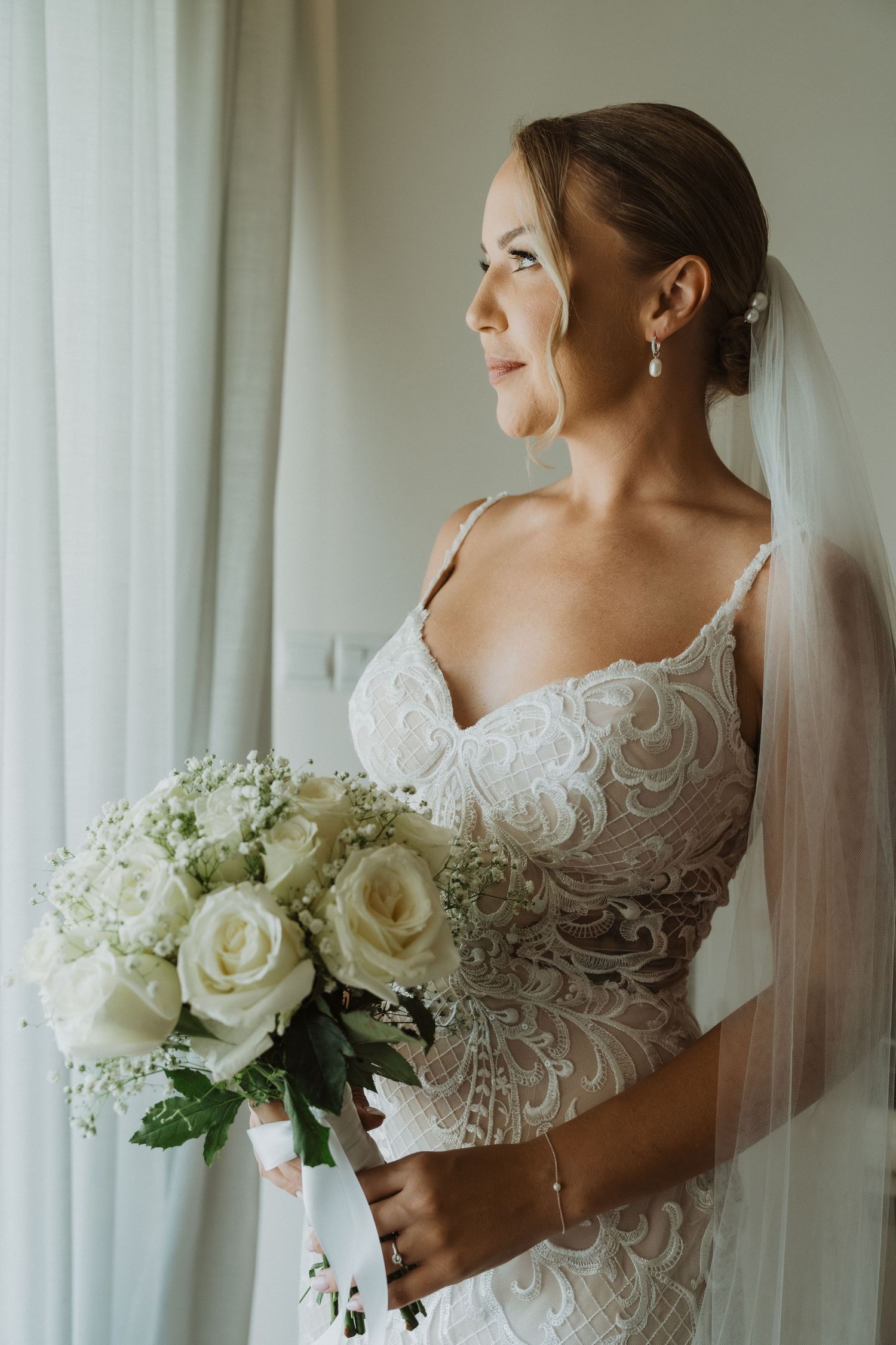 Bride in lace wedding dress holds elegant white rose bouquet while standing by window in soft natural light.