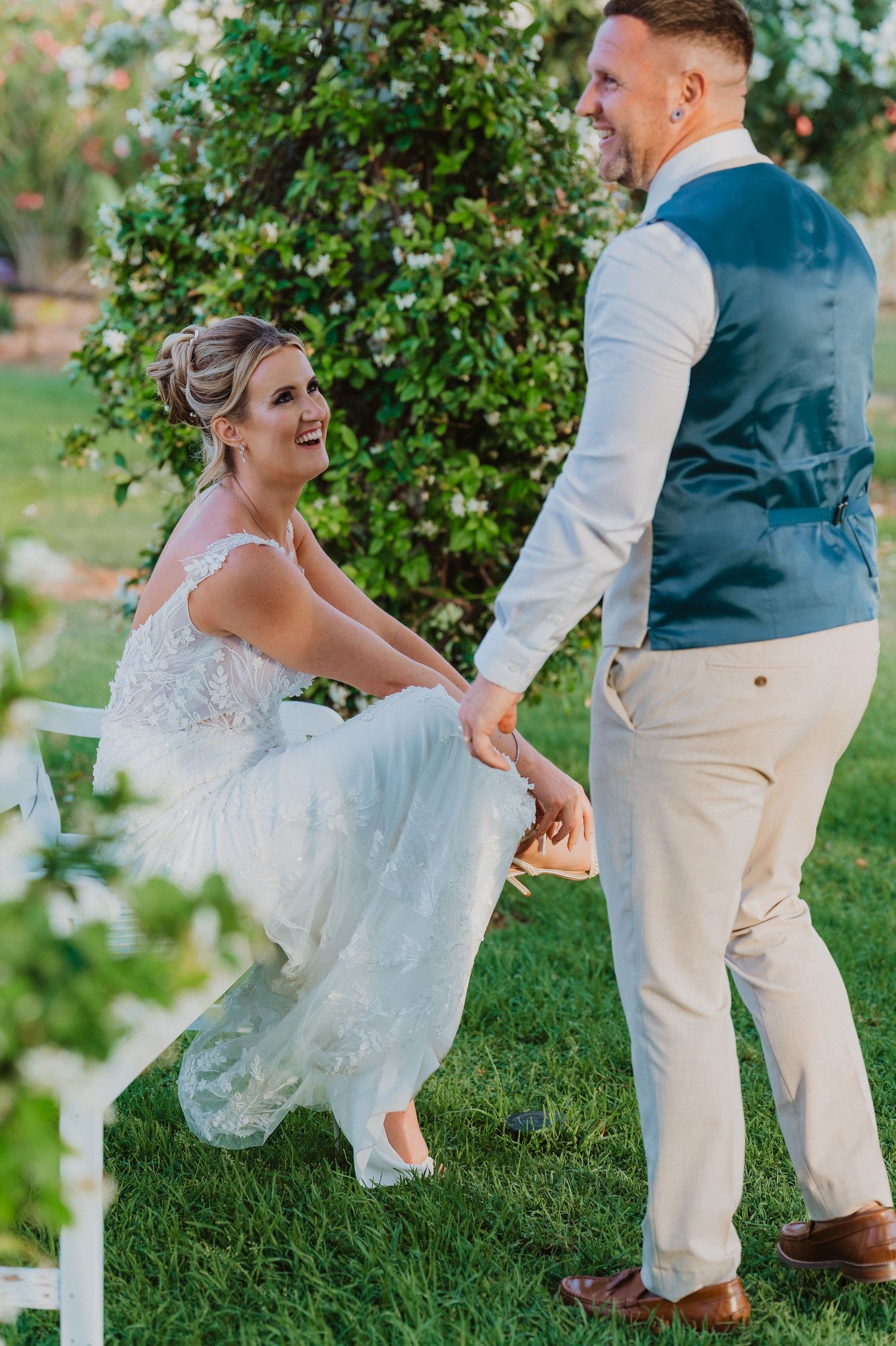 A couple shares a playful moment in a garden setting as the bride lifts her lace dress to show her shoes.