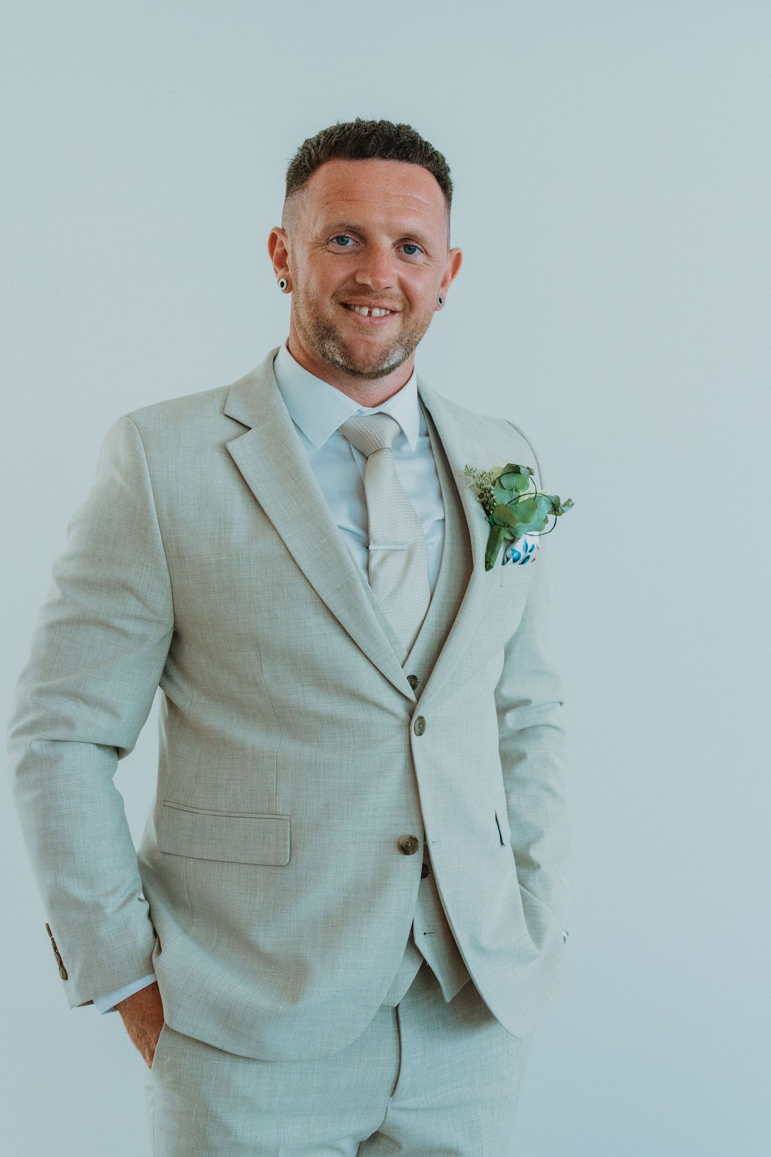 A person in a light beige suit poses against a pale blue background with a boutonniere.