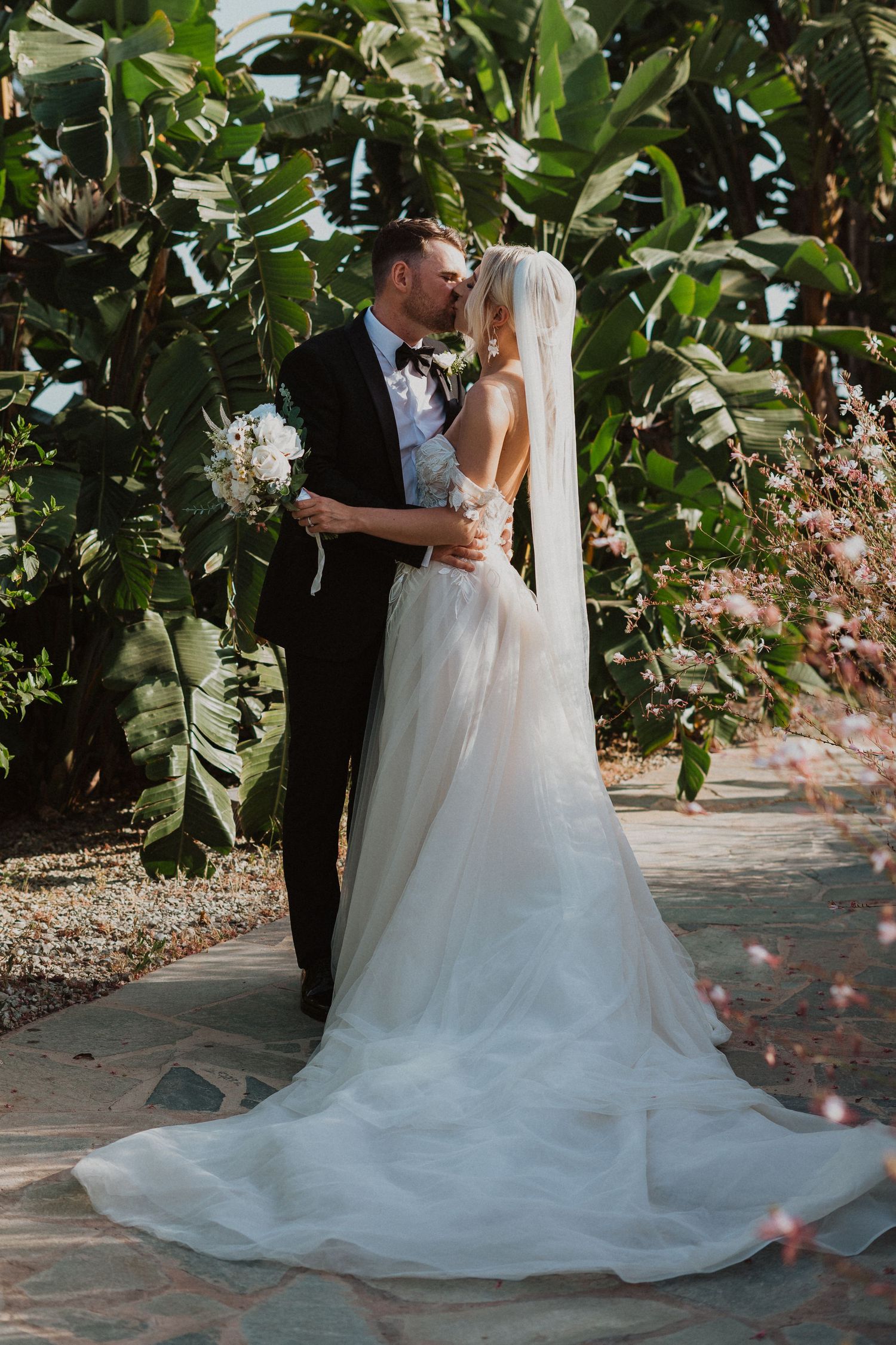 A wedding couple shares a tender moment on a stone path surrounded by tropical foliage.