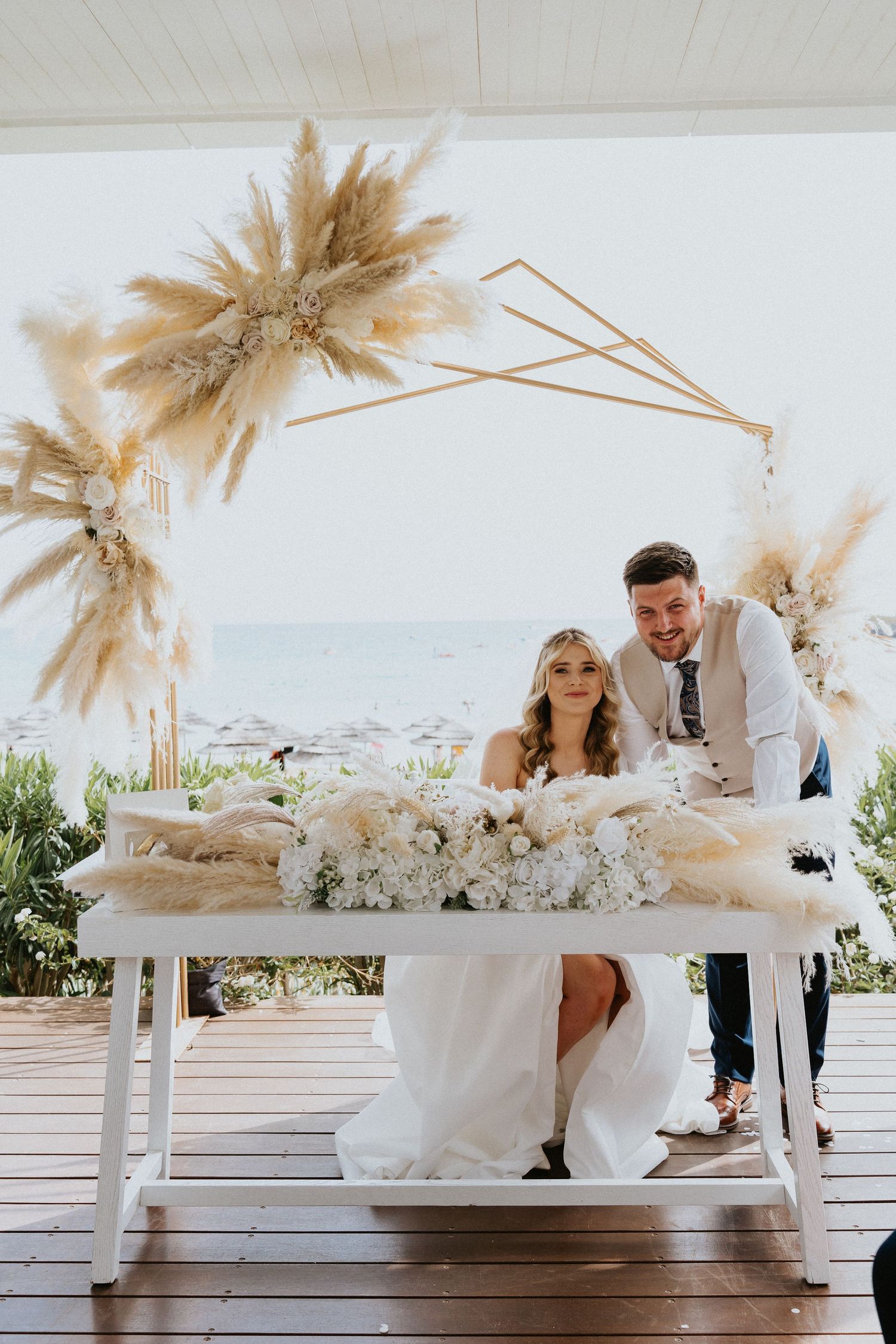 Wedding couple poses at decorated dessert table with pampas grass arch and ocean view.