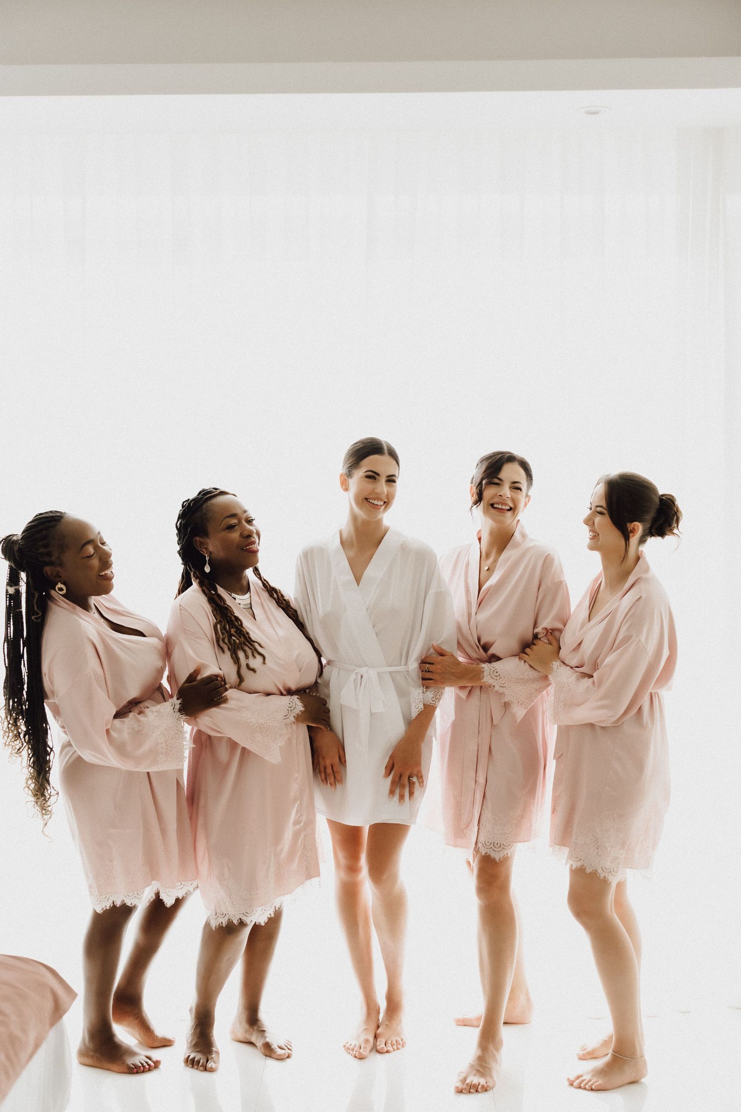 Bridesmaids in blush robes gather for a pre-wedding photo against a white background.