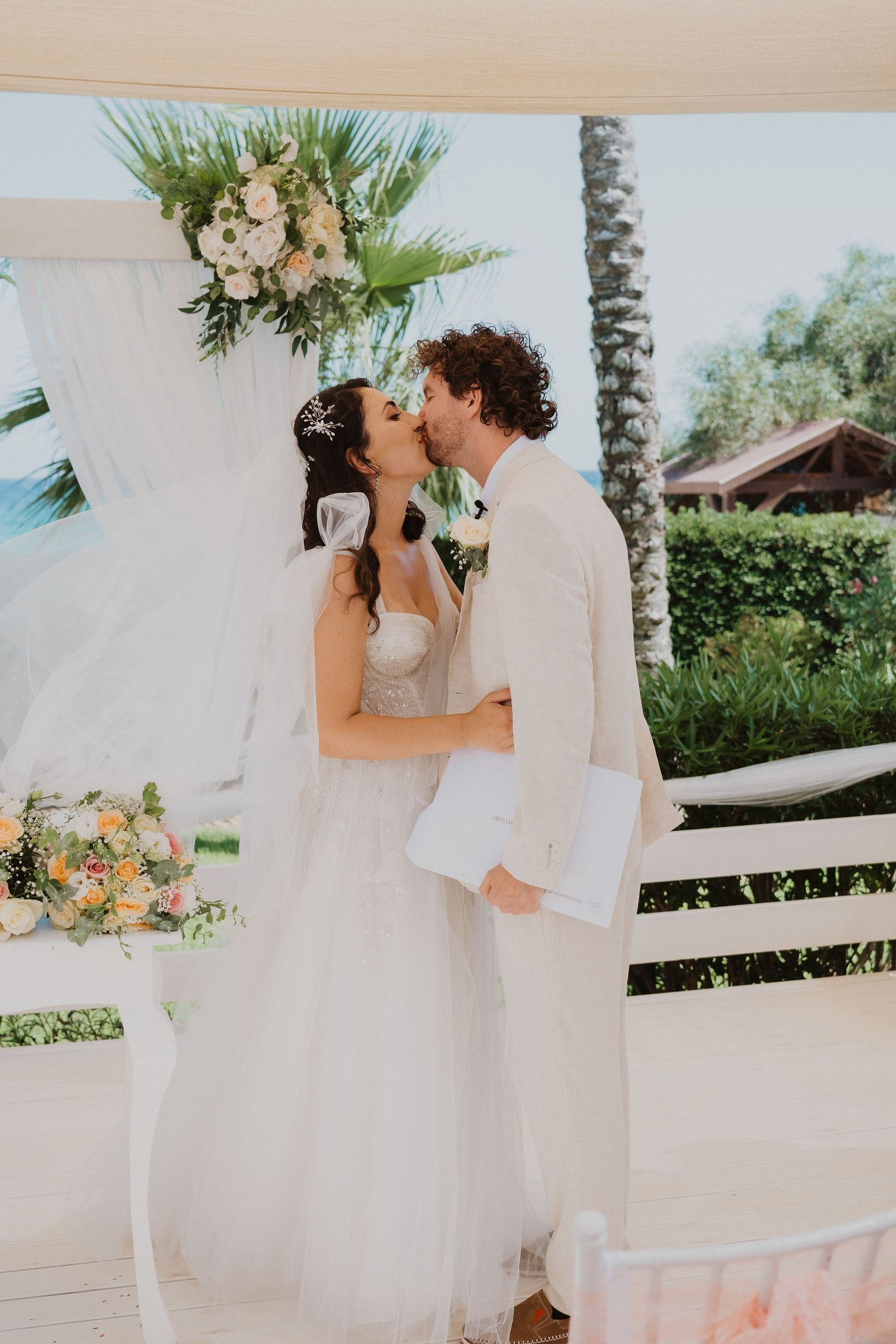 Romantic wedding kiss under floral decorated arch with tropical beach setting.