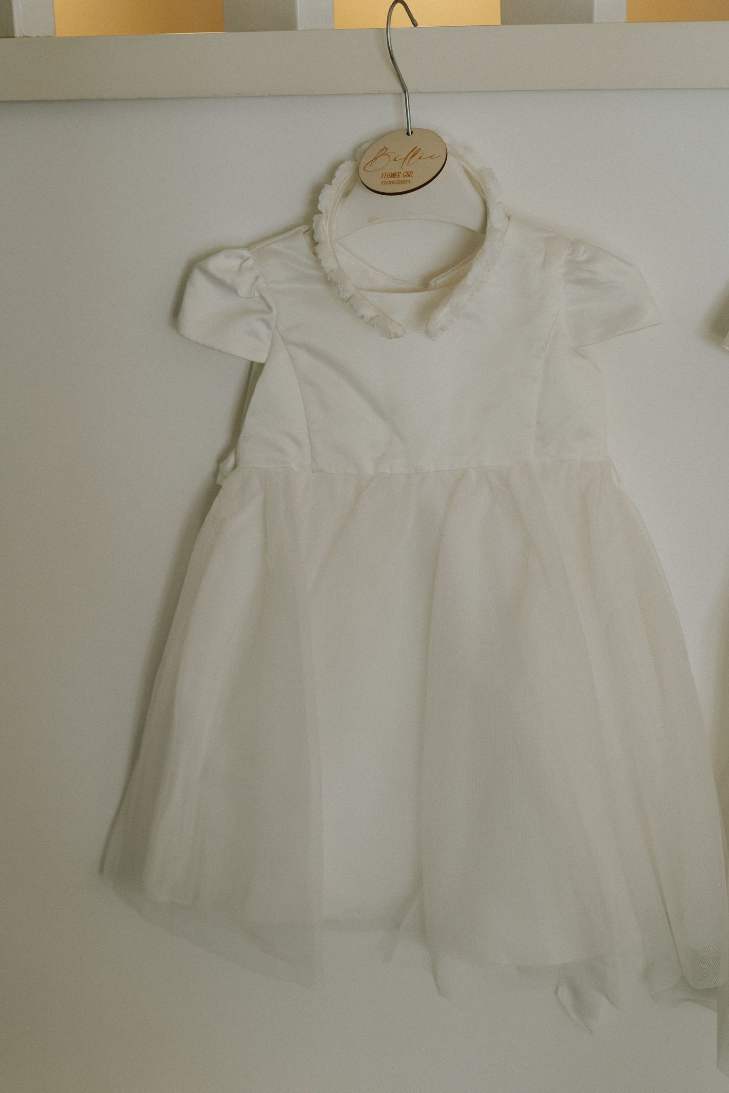 White flower girl dress with puff sleeves hanging on a wooden hanger against grey wall.