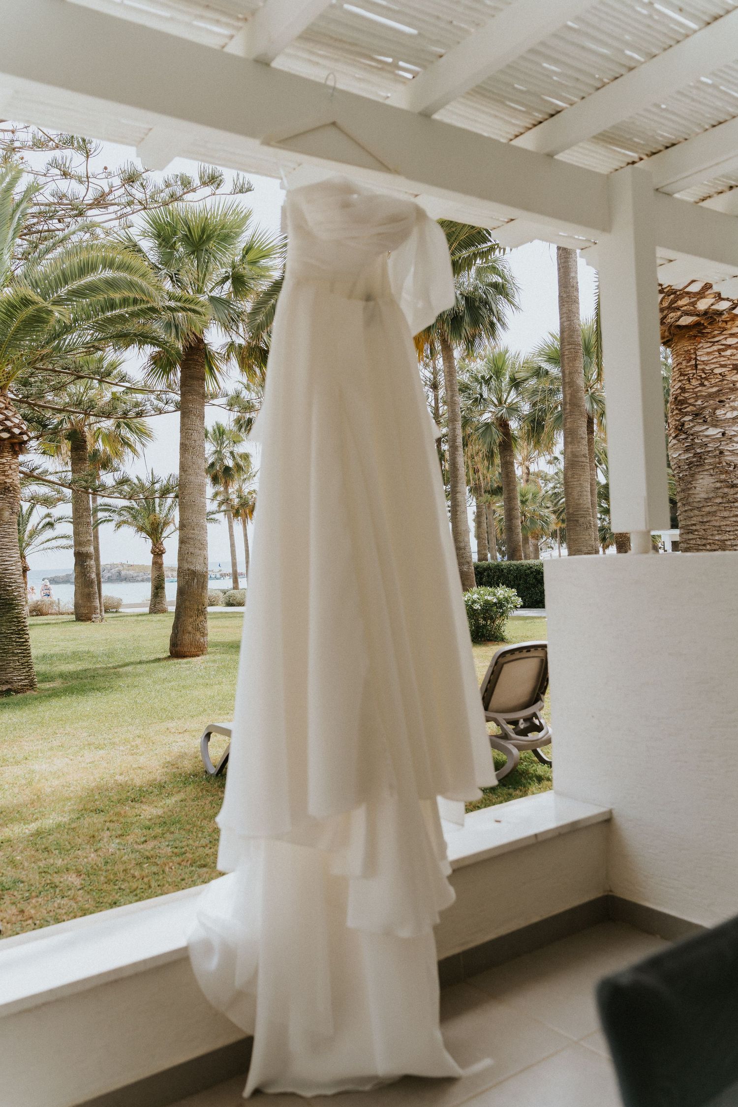 Flowing white wedding dress hanging on pergola with tropical palm trees in background.
