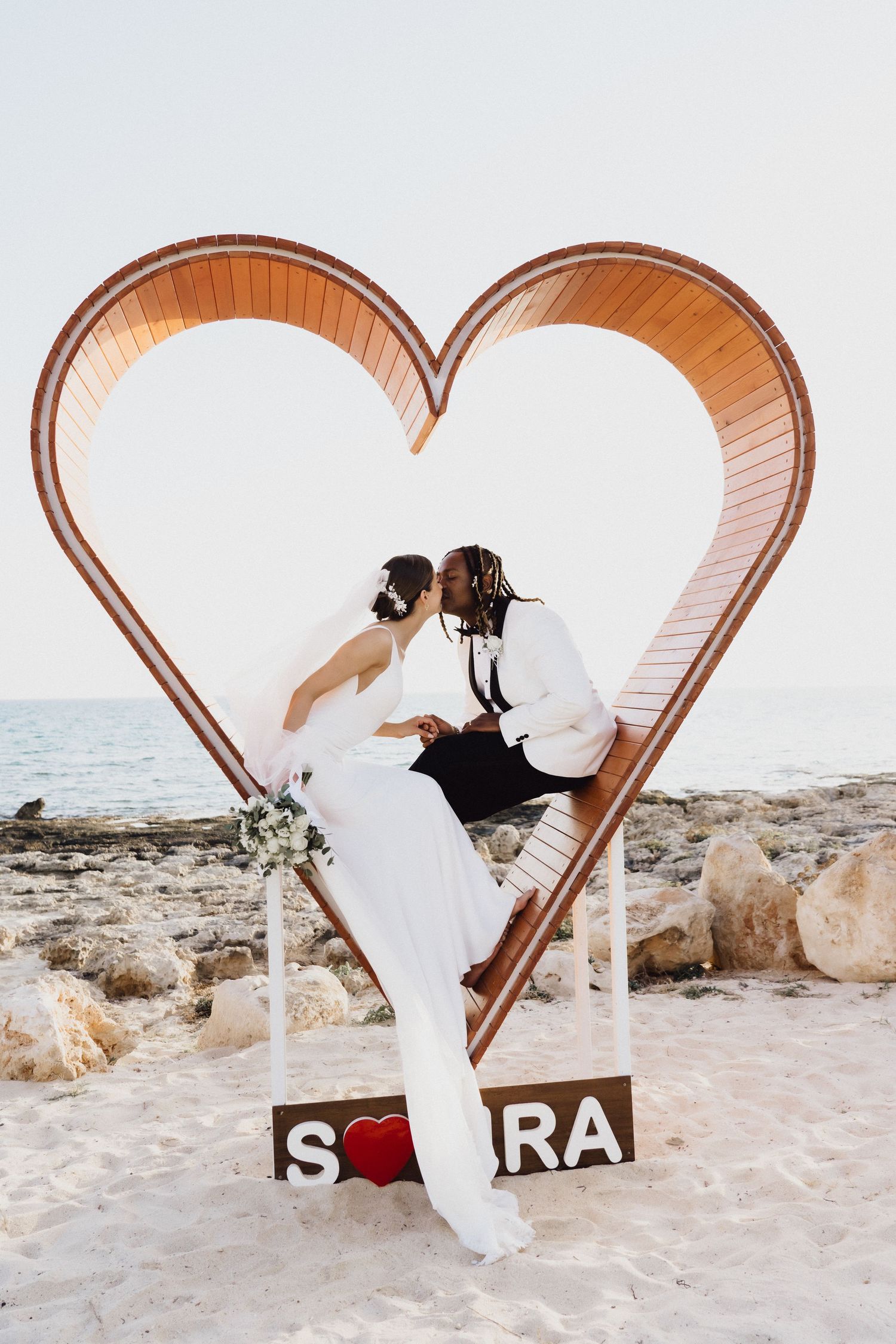 Newlywed couple embraces inside a rustic wooden heart frame display on a beach at sunset.