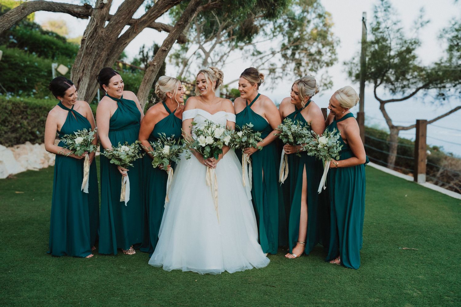 Wedding party in emerald green bridesmaid dresses poses on a grassy hillside overlooking the ocean.