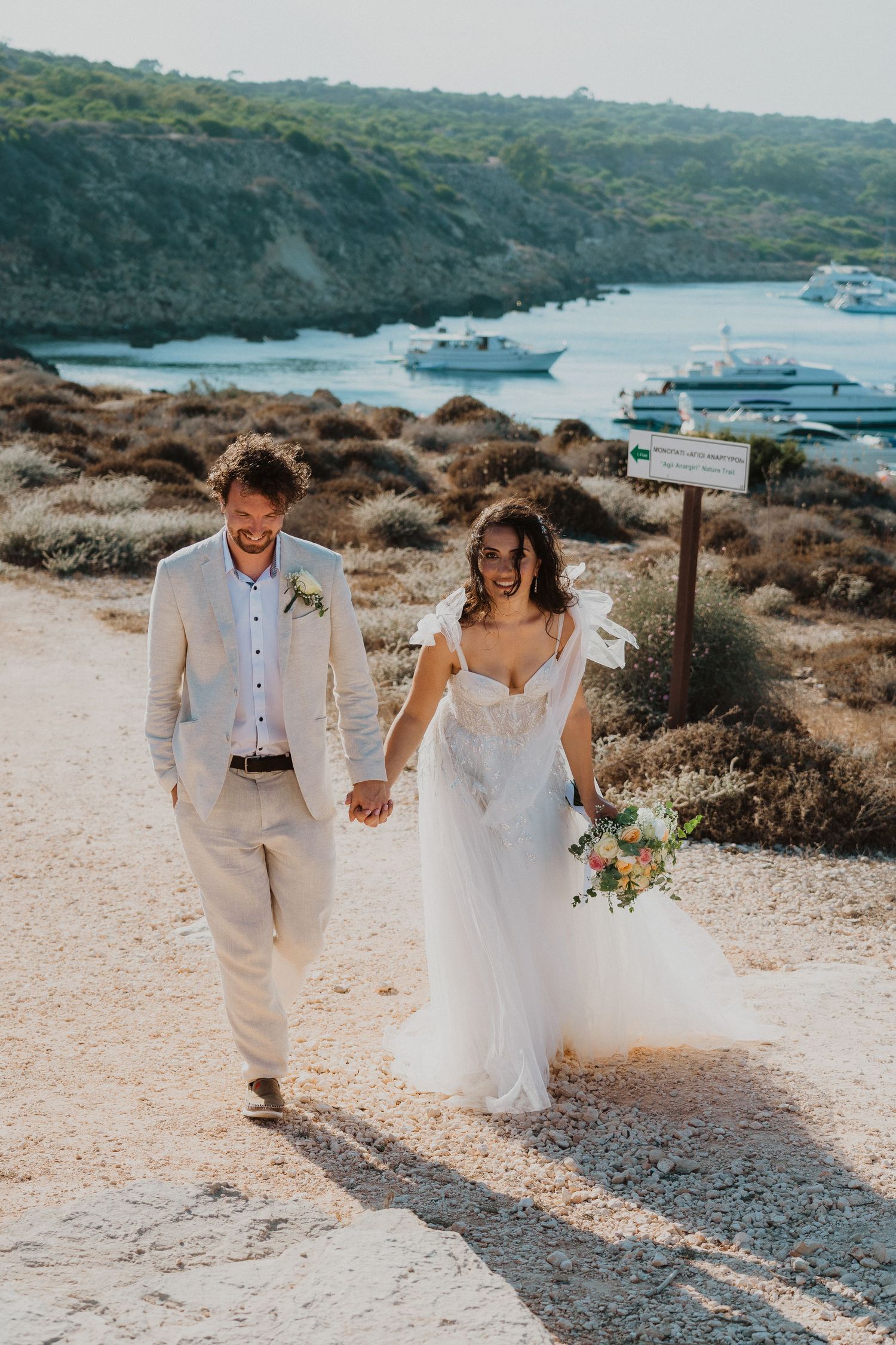 A couple holding hands during their seaside wedding ceremony with boats in the bay along a rugged Mediterranean coastline.