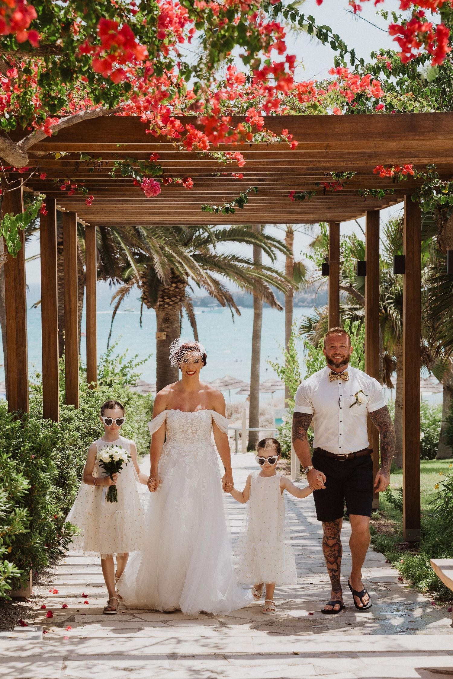 Family strolls under a wooden pergola decorated with vibrant red bougainvillea flowers.