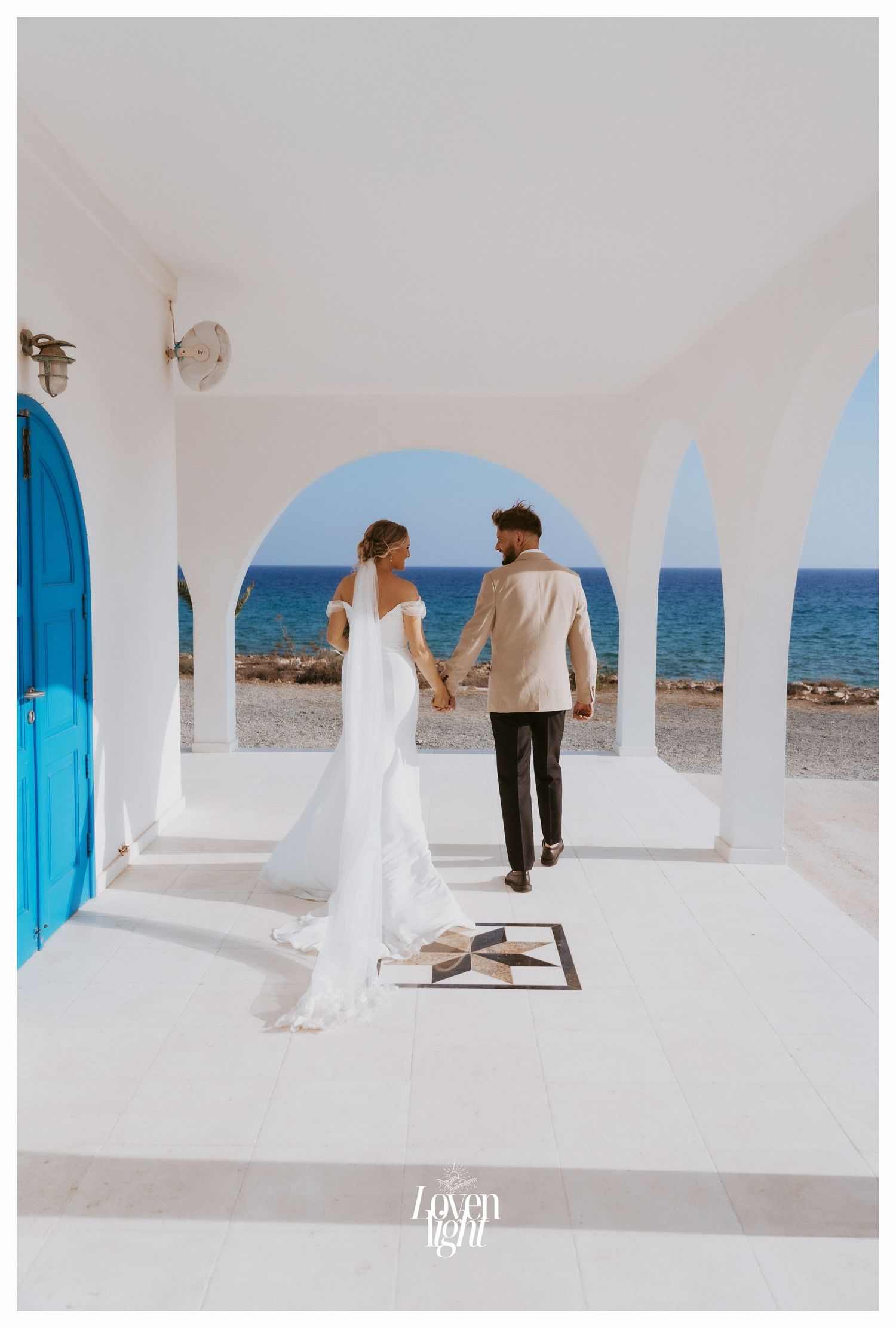 Newlyweds holding hands while walking under Mediterranean white arches with turquoise door and seaside backdrop.