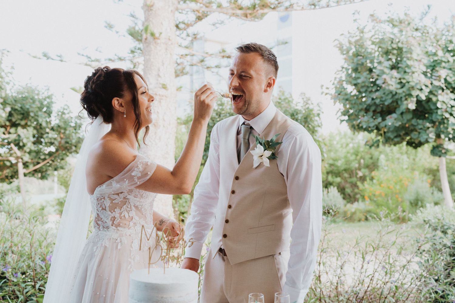 A romantic outdoor wedding moment where the bride playfully feeds cake to the groom in a garden setting.