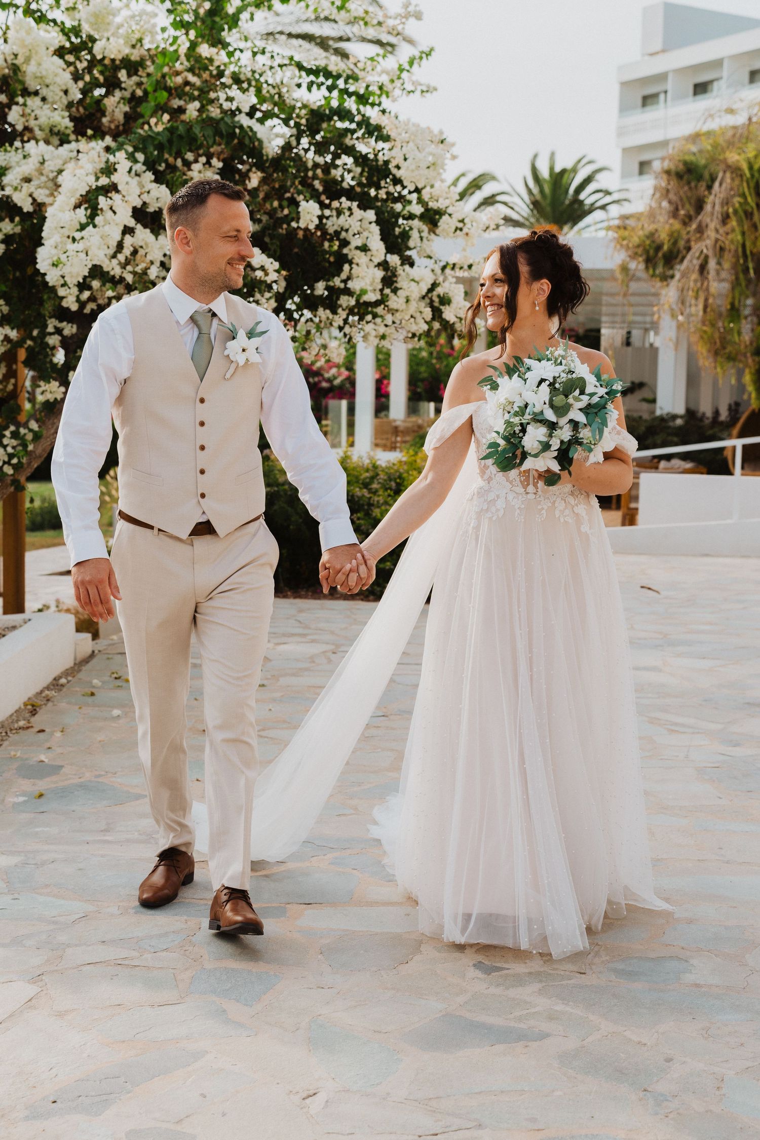 A newlywed couple walks hand in hand on a stone path with blooming white flowers and palm trees in the background.