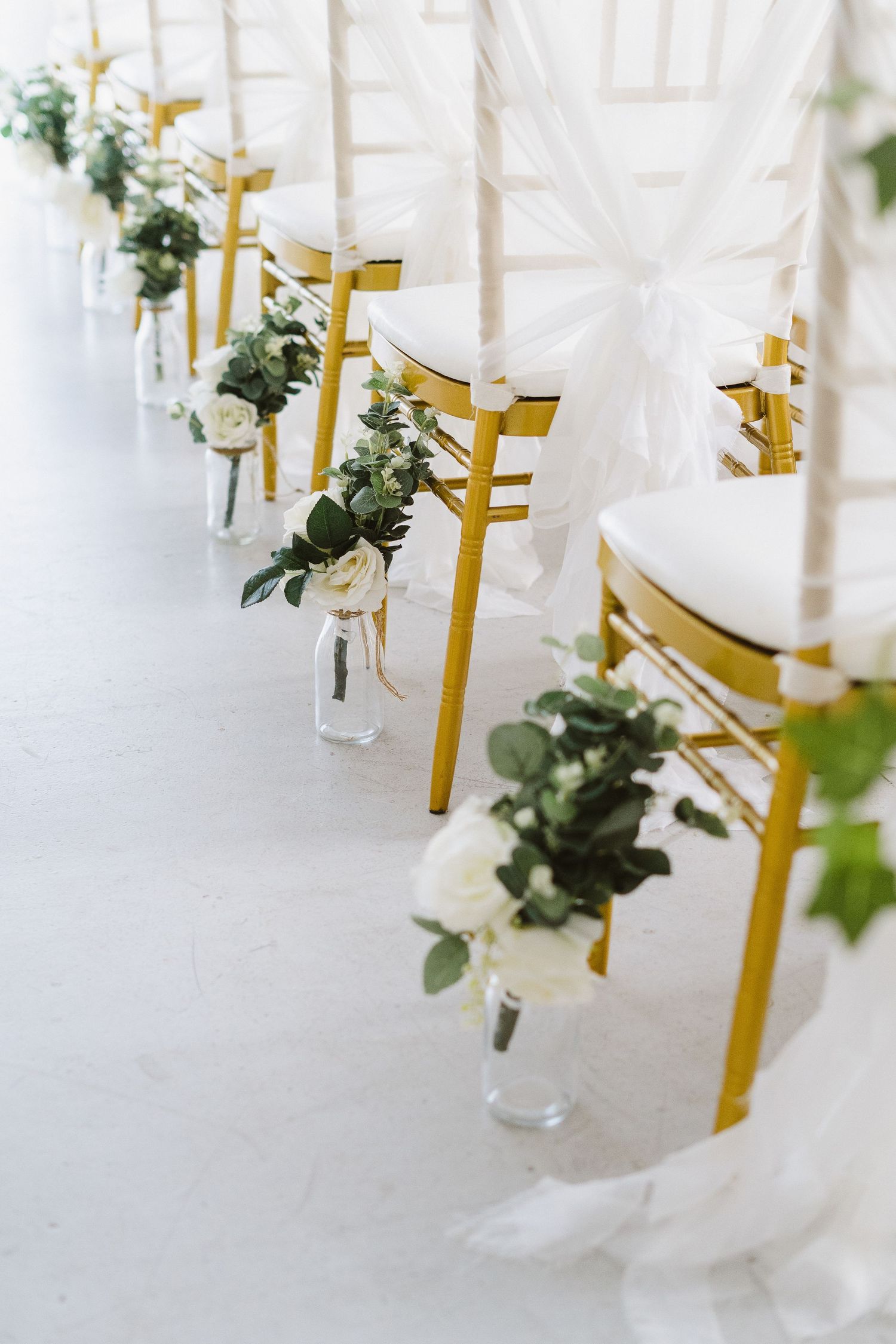 Row of gold chiavari chairs with white floral arrangements hanging in glass vases.