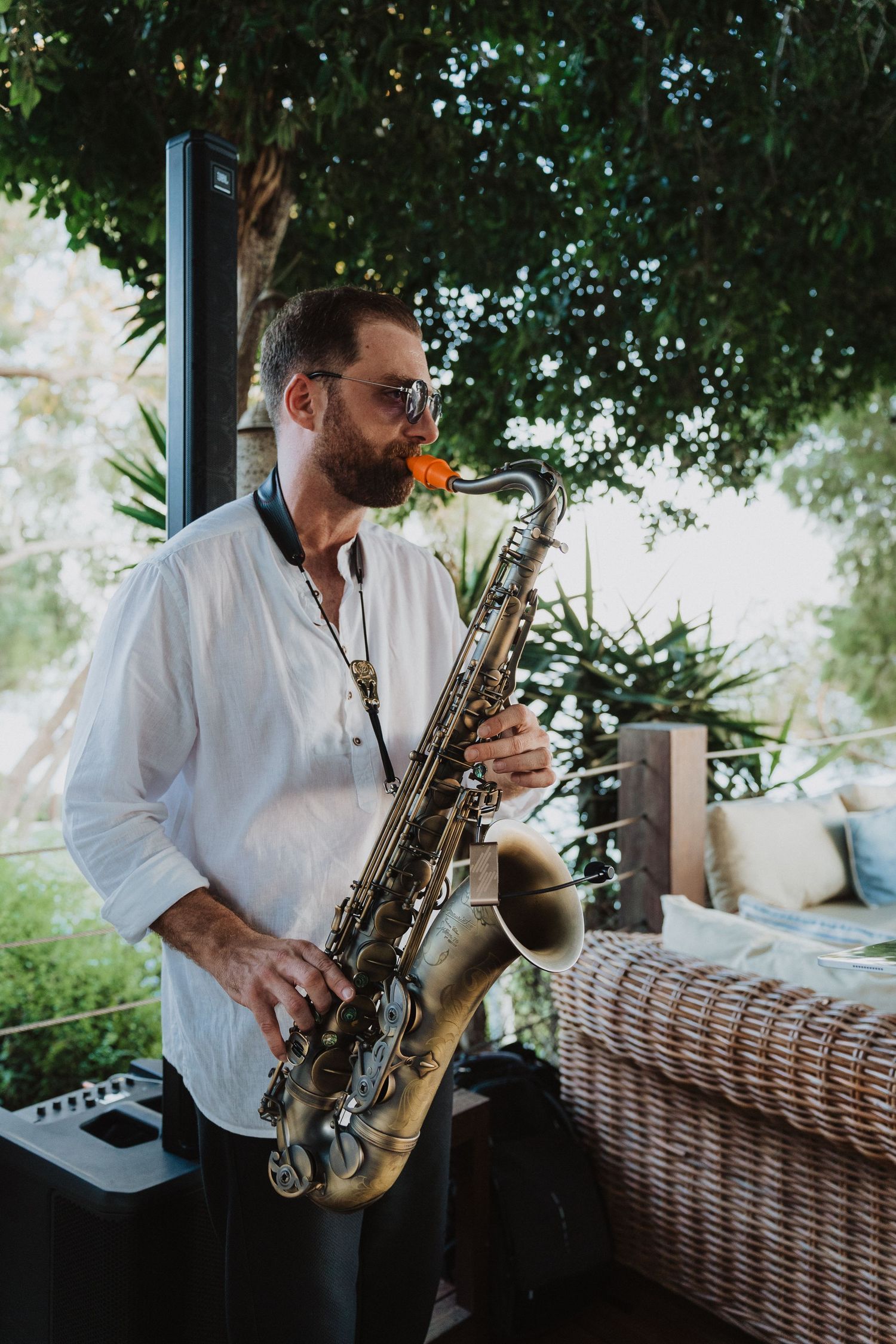 Musician in white button-down shirt playing saxophone on outdoor patio with rattan furniture and greenery.