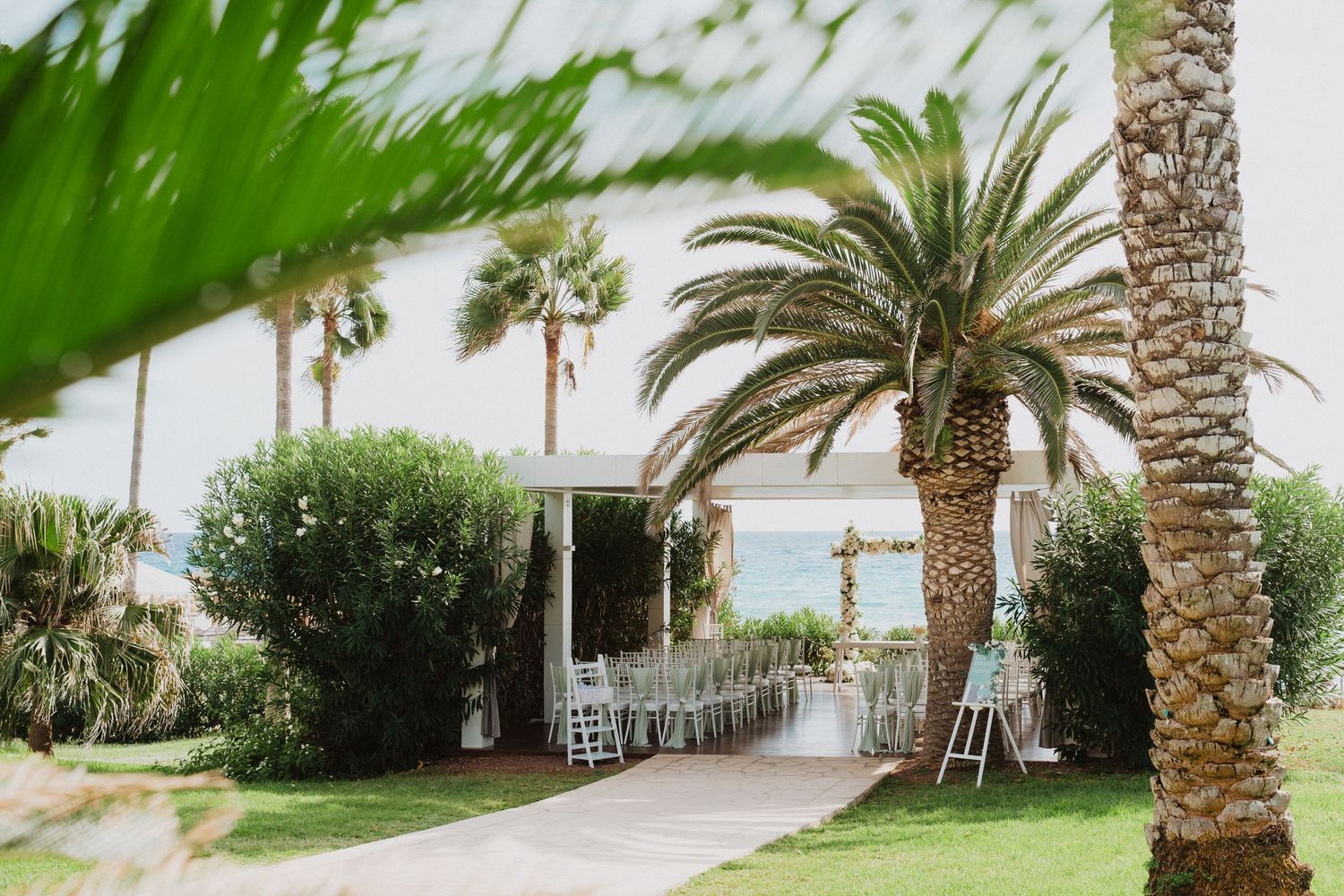 An elegant outdoor wedding venue pathway lined with palm trees and green foliage leading to an ocean view.