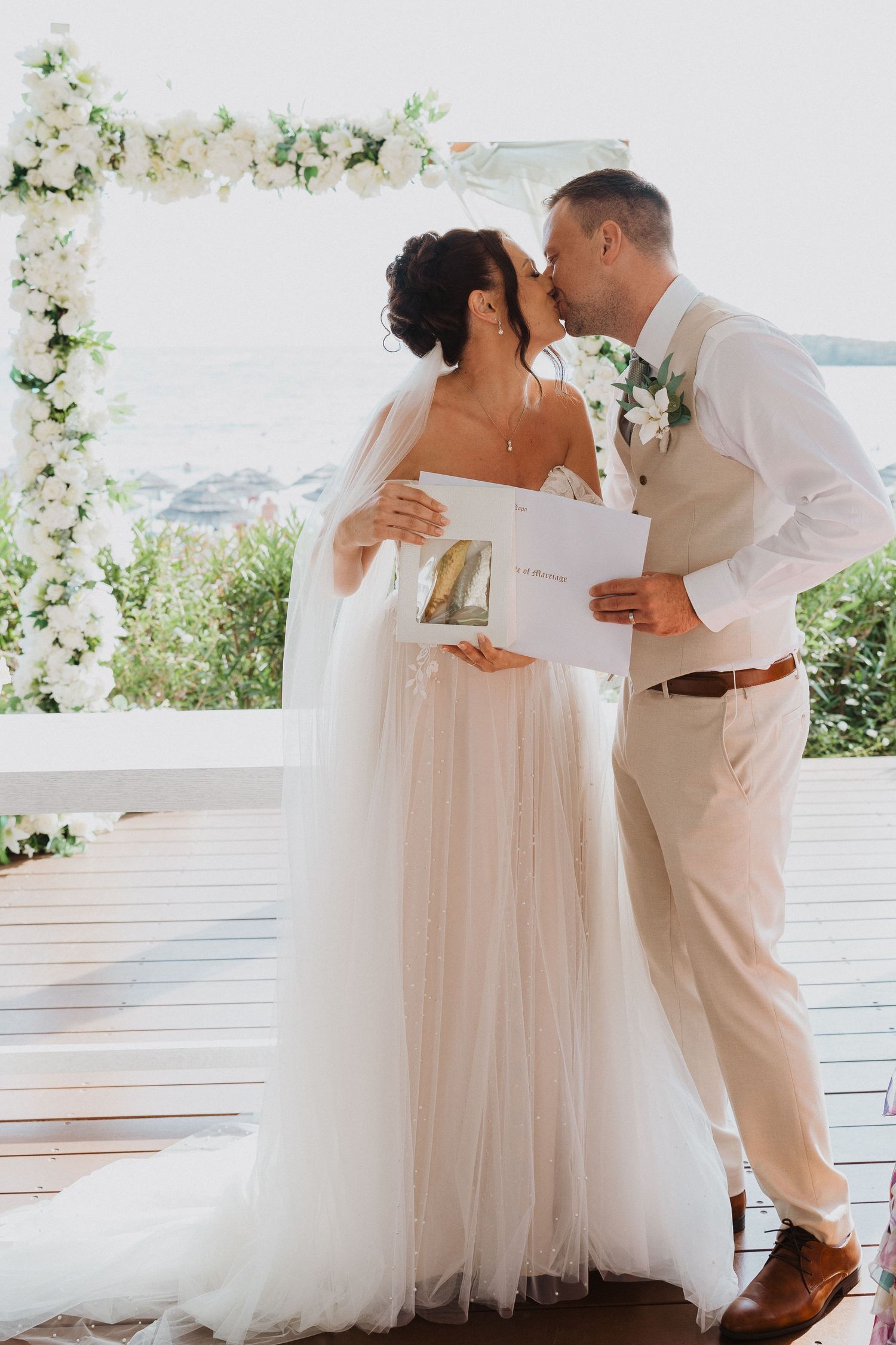 A romantic moment captured at a seaside wedding ceremony under a white floral arch on a wooden deck.