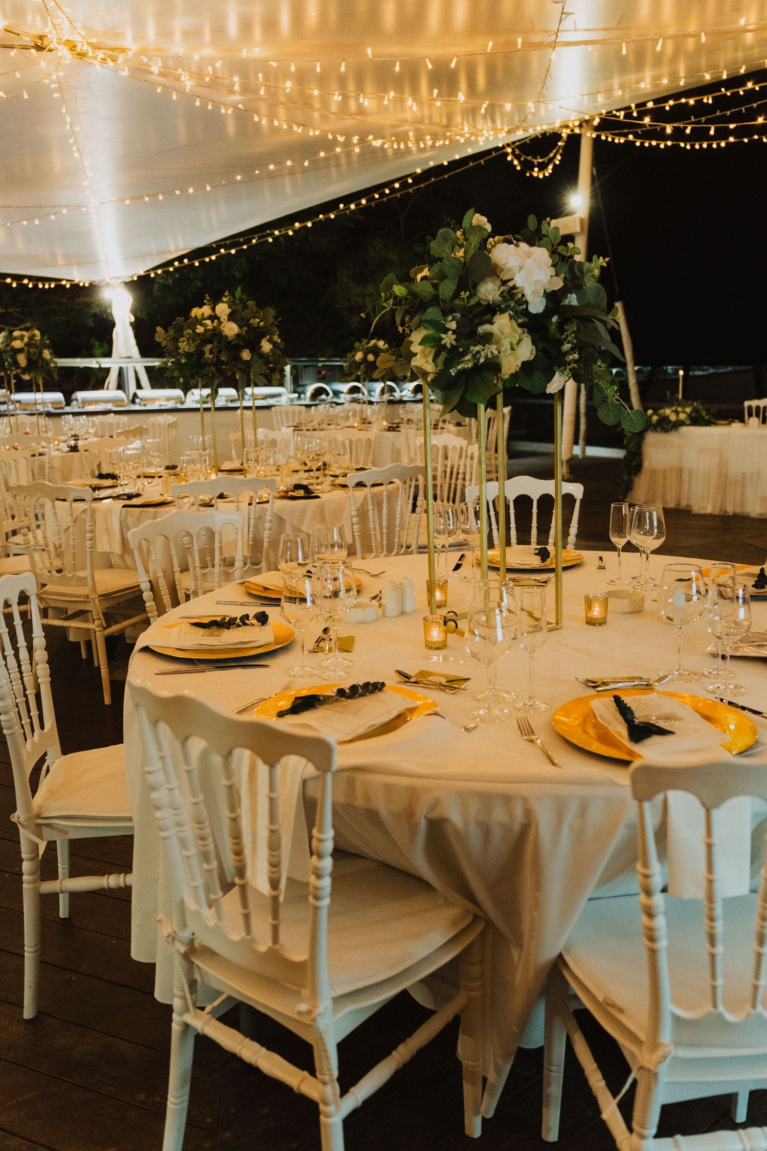 Formal wedding reception table setting with white linens, gold chargers and elegant floral centerpieces under string lights.