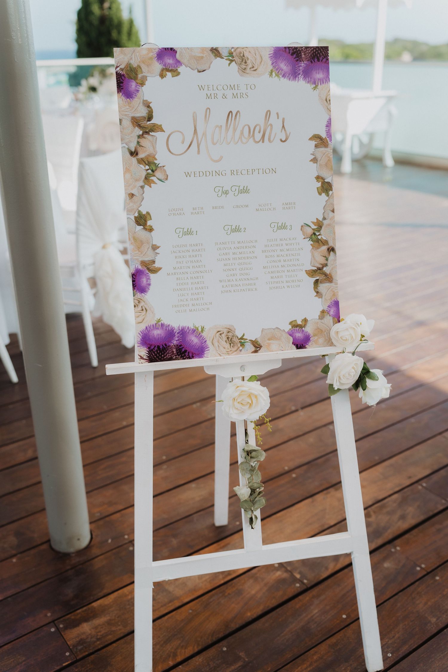 Wedding seating chart display with purple and gold floral design on white wooden easel.