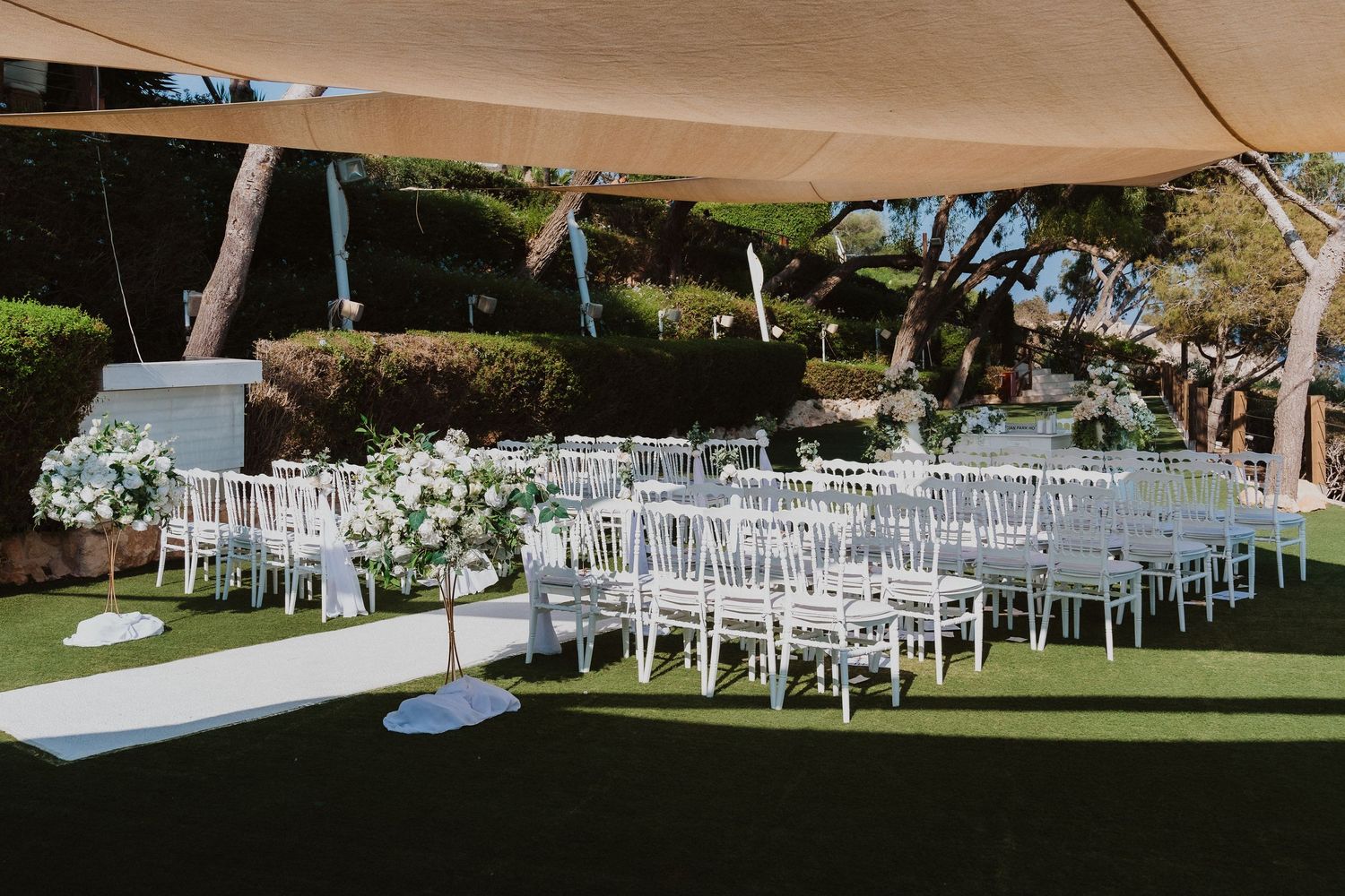 Elegant outdoor wedding ceremony setup with white chairs and floral arrangements under a beige canopy on a grassy lawn.