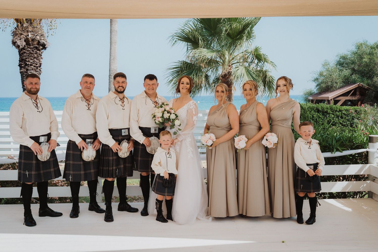 A wedding party in formal attire poses outdoors with bridesmaids in beige dresses and groomsmen in kilts by the ocean.