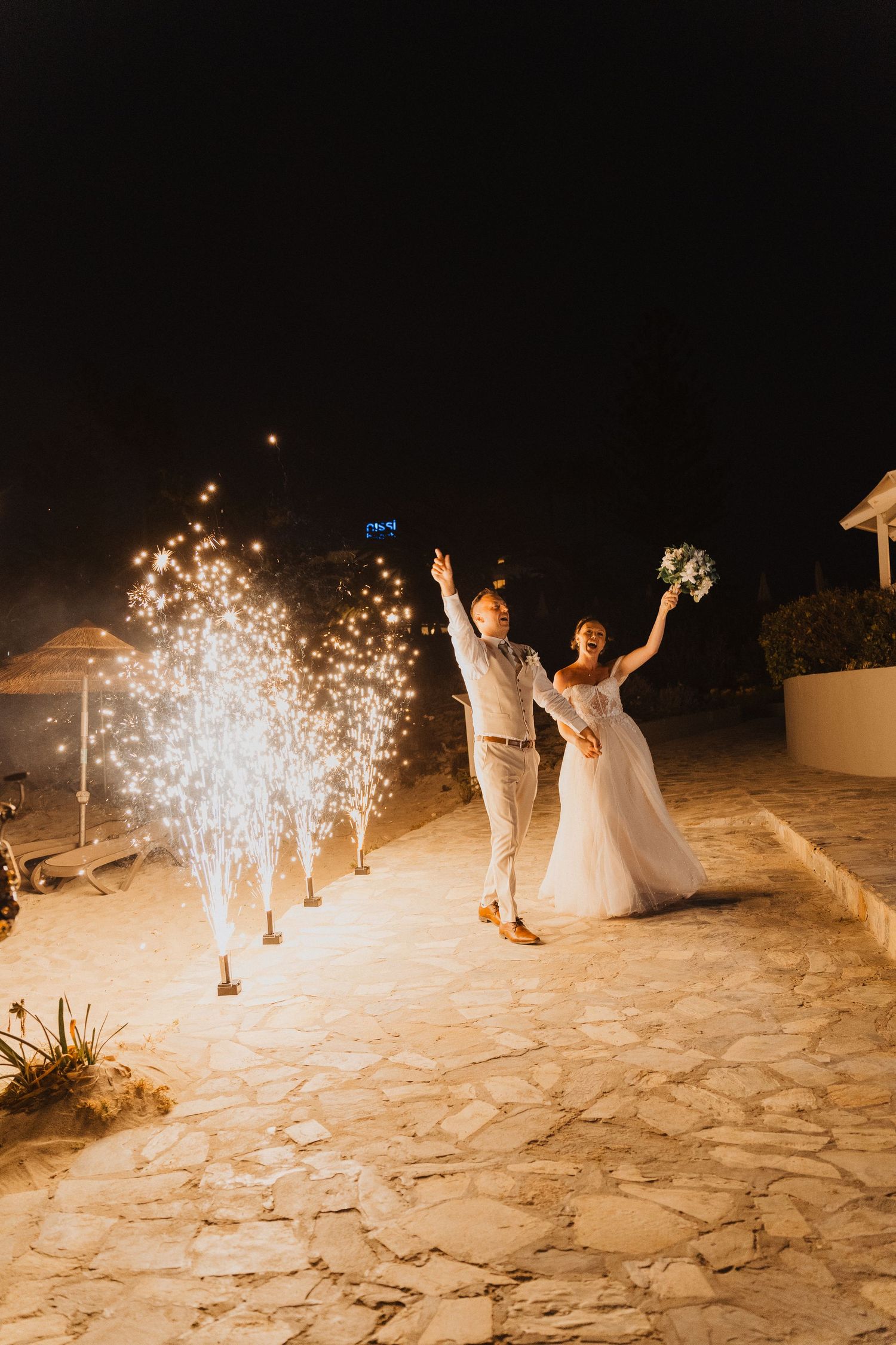 Newlyweds celebrate on stone patio at night with sparklers creating magical fountains of light in background.