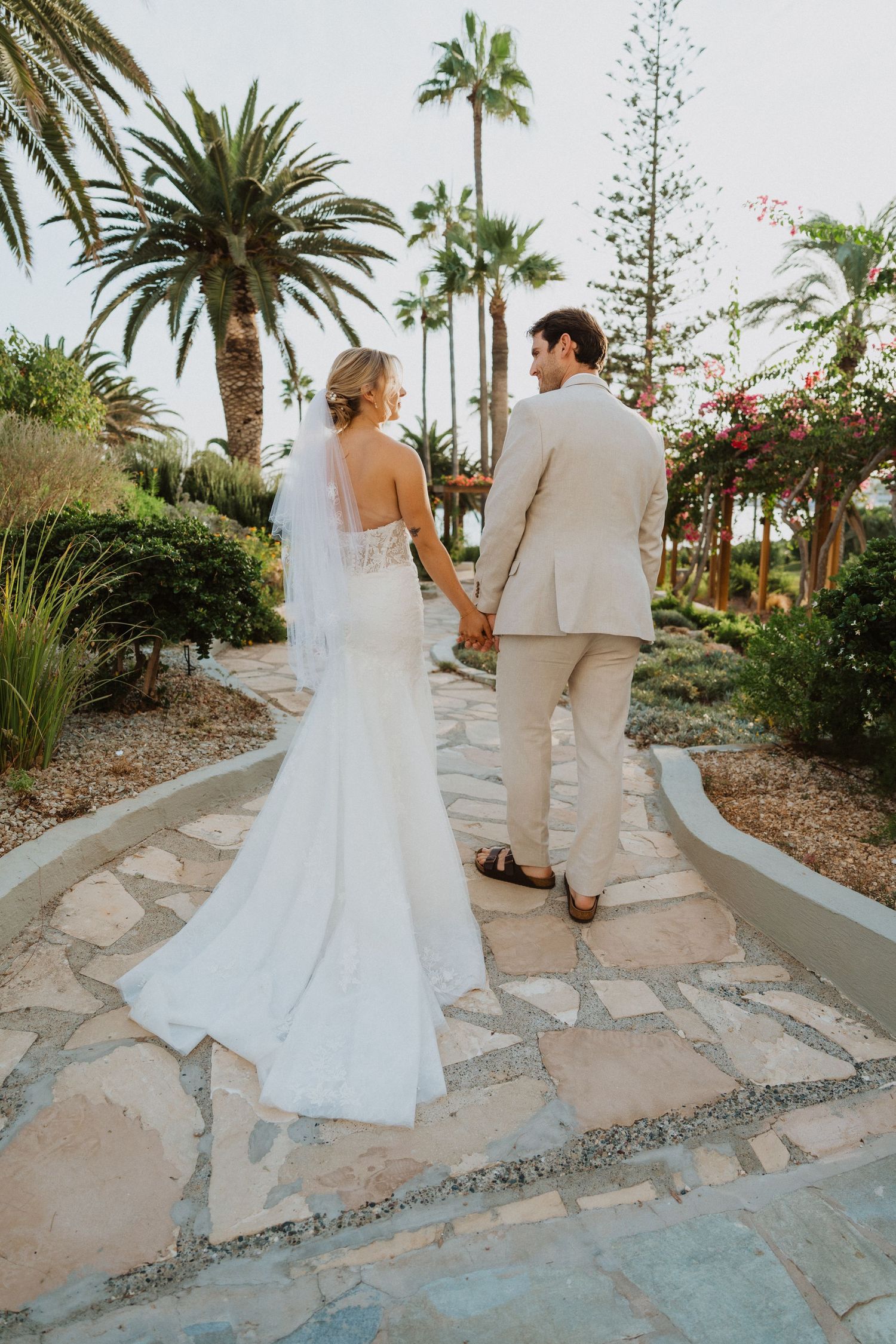 Newlyweds walk along a winding stone path surrounded by tropical palm trees and flowers.