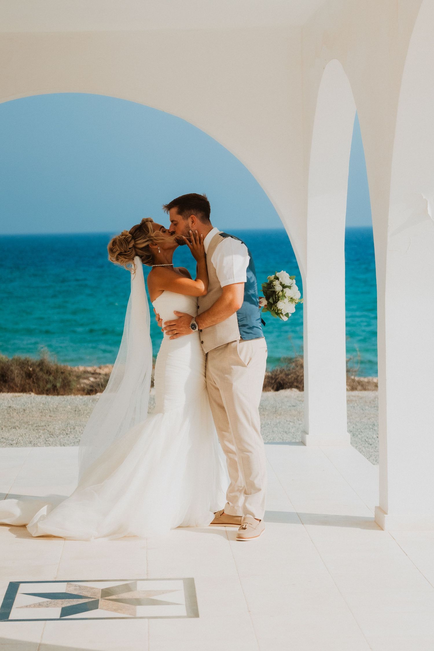 Bride and groom kissing under white arches with turquoise ocean backdrop at a romantic beach wedding ceremony.