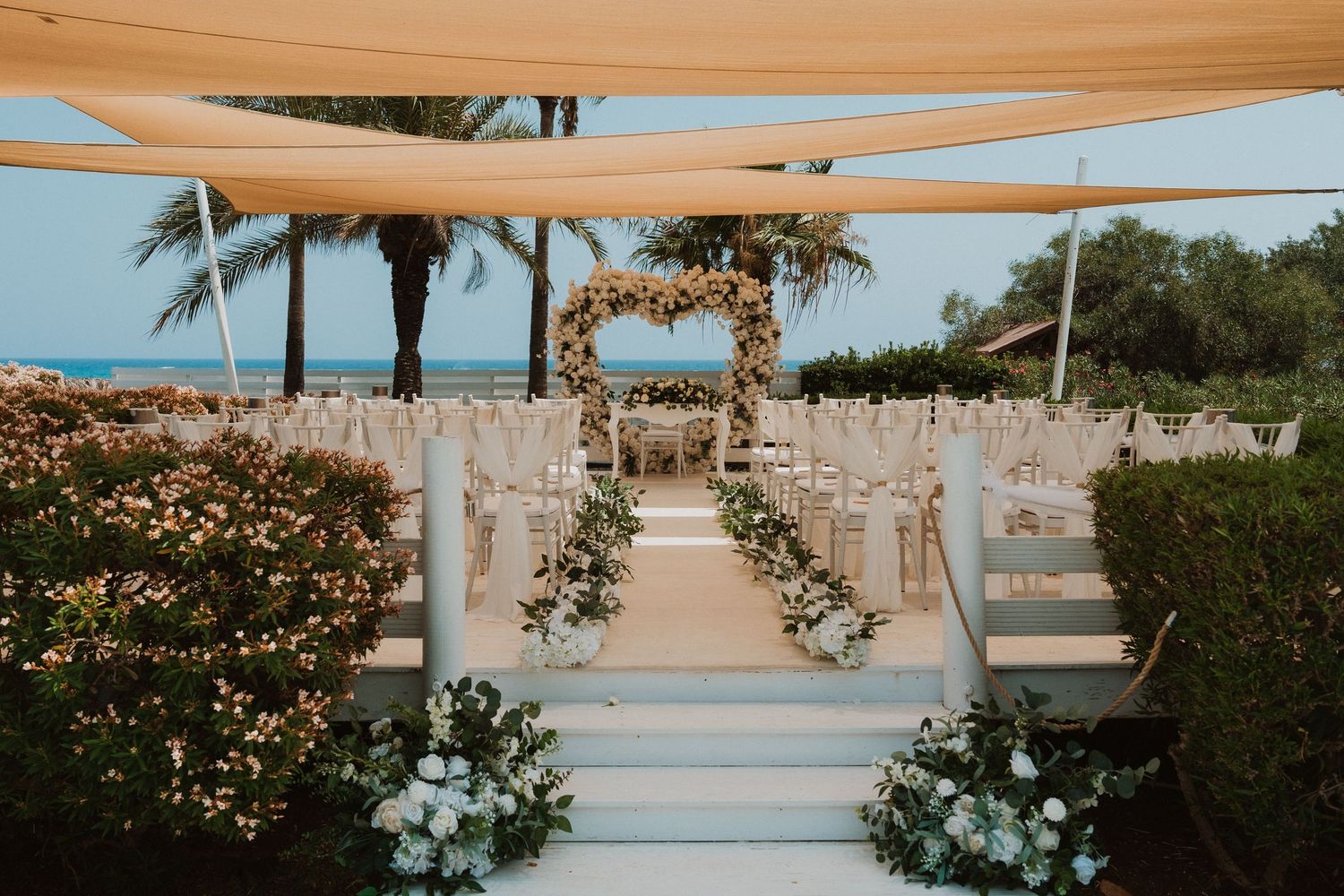 Elegant outdoor beach wedding ceremony setup with floral arch, white chairs, and lush greenery under a canopy.
