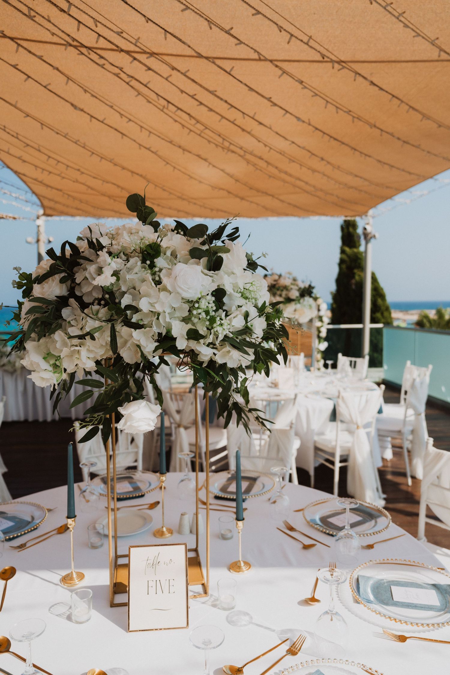Elegant outdoor wedding reception table with gold centerpiece, white floral arrangement, blue candles, and gold cutlery under string lights.