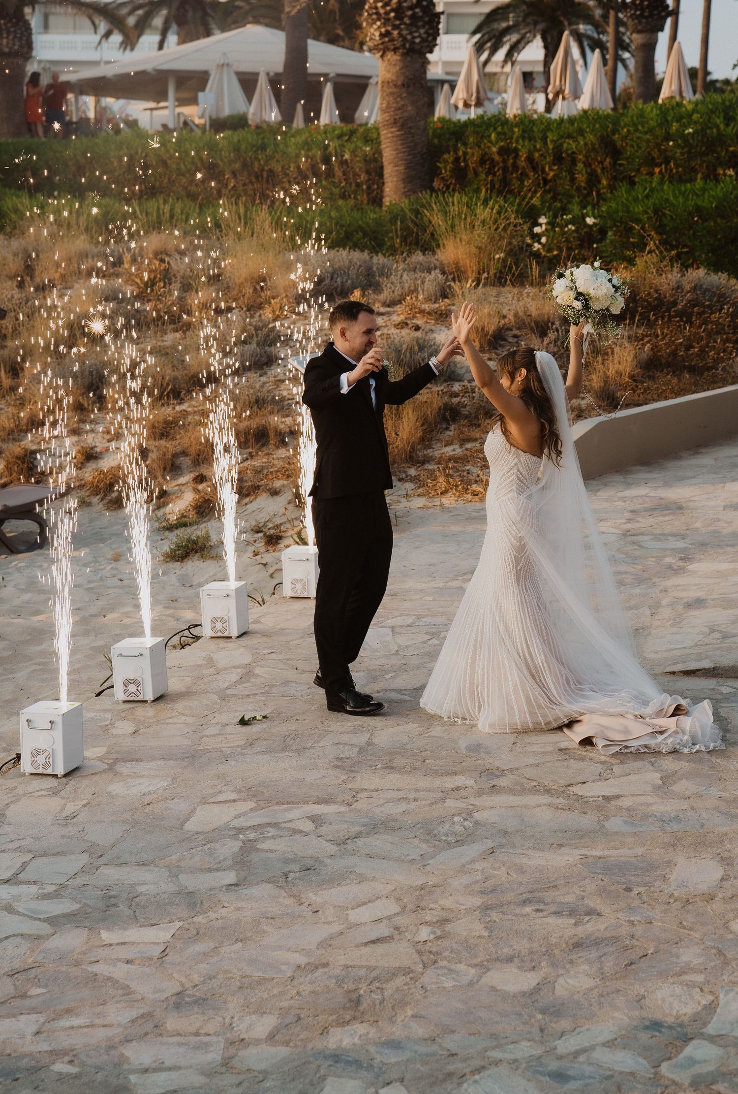 Bride and groom celebrating with fountain sparklers on a stone patio surrounded by palm trees at golden hour.