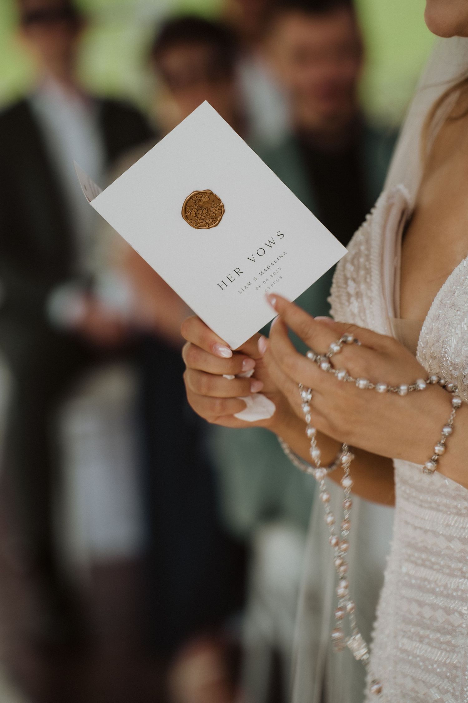 Bride holding a white wedding vow booklet with gold wax seal, wearing pearl bracelet and beaded dress.