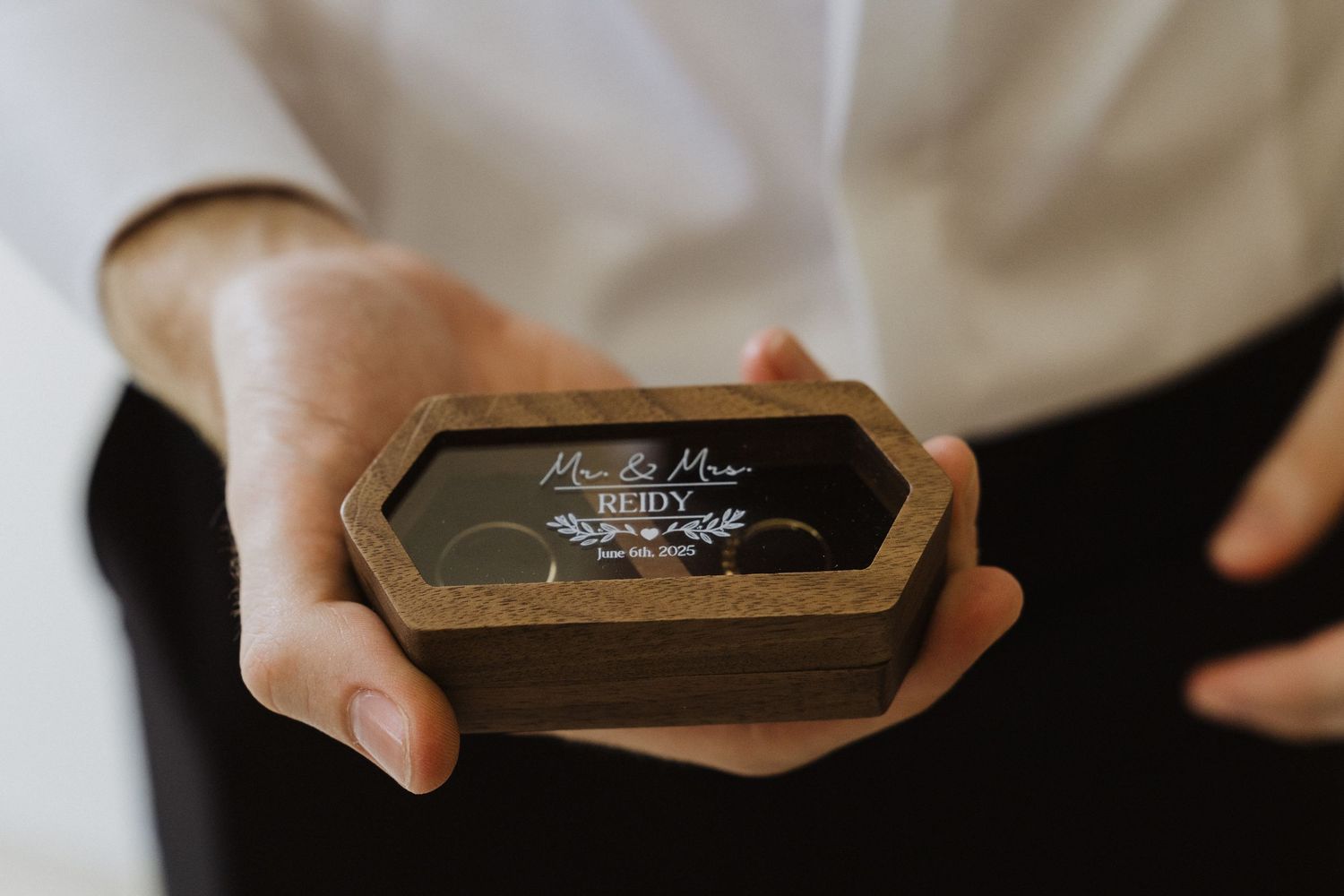 Groom holding a wooden hexagonal ring box with personalized engraving displaying wedding rings inside.