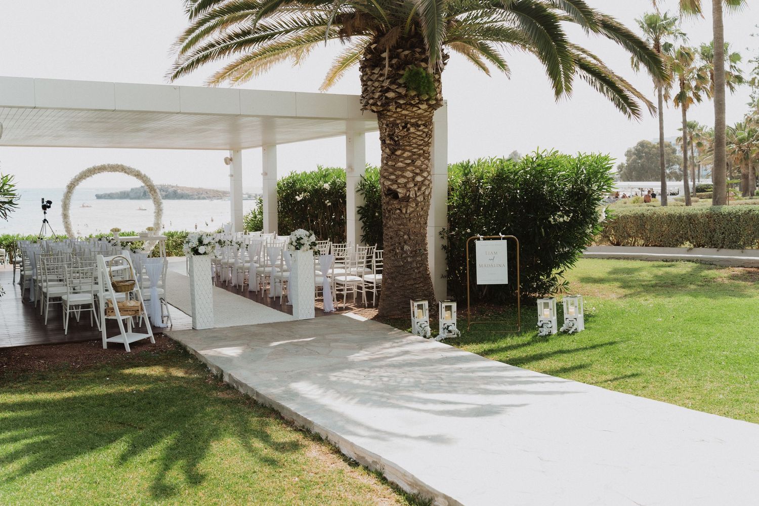Outdoor wedding ceremony setup with white chairs under pavilion surrounded by palm trees.