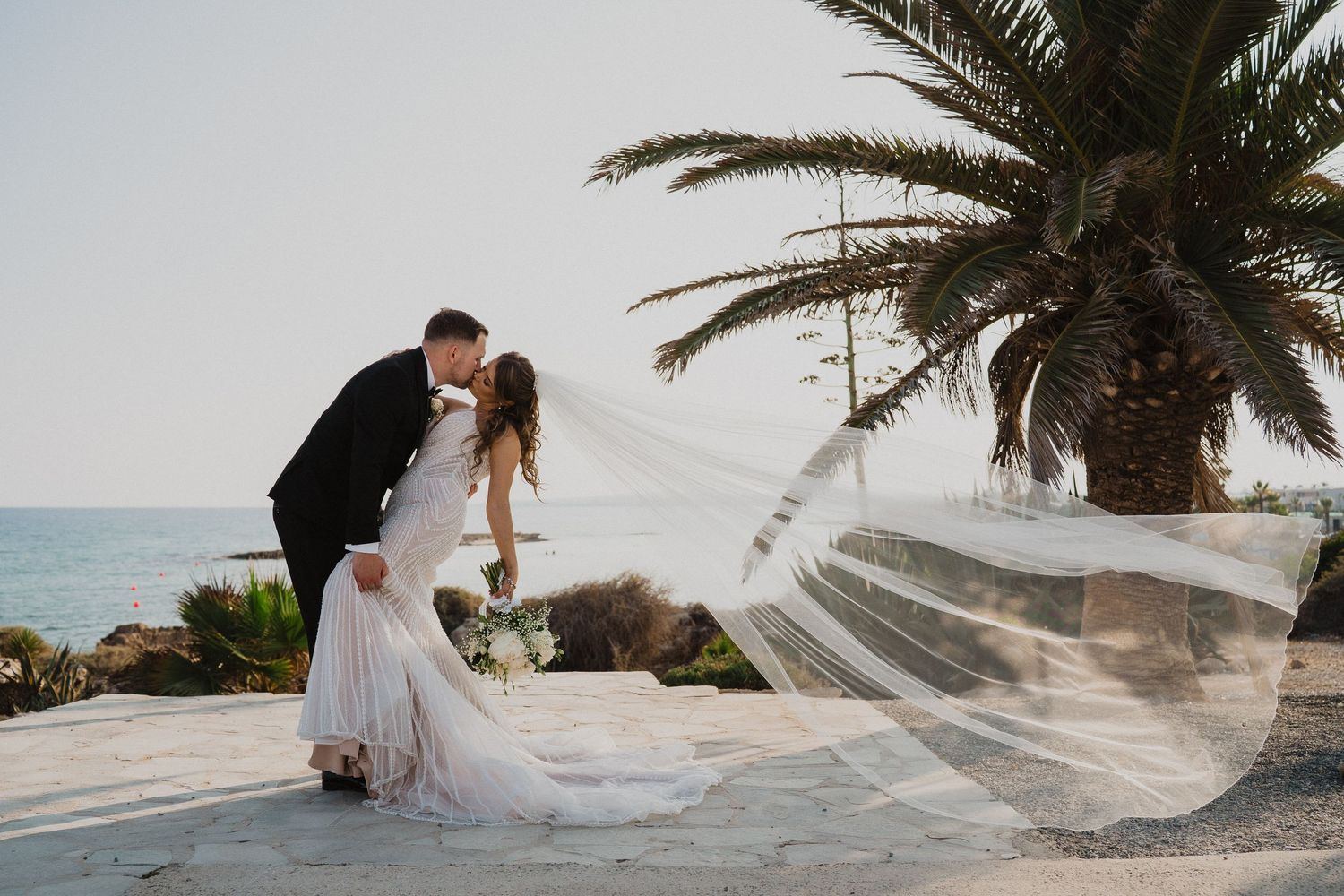 Bride and groom share a dip kiss near a palm tree with flowing veil and ocean backdrop on wedding day.