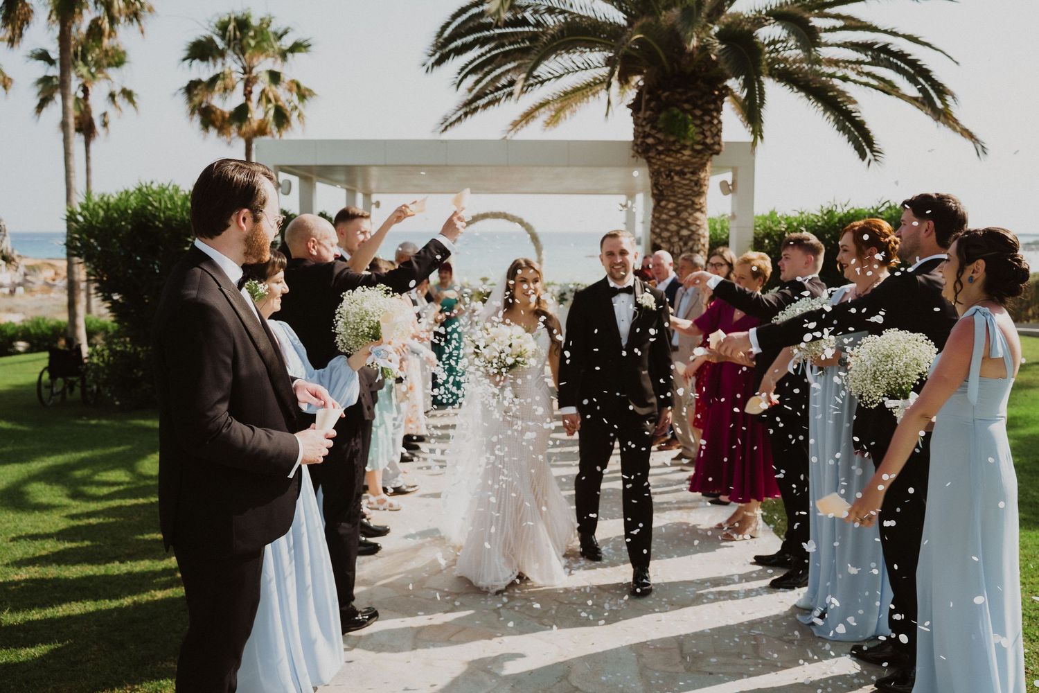 Bride and groom walk through confetti shower surrounded by cheering guests at outdoor wedding ceremony.