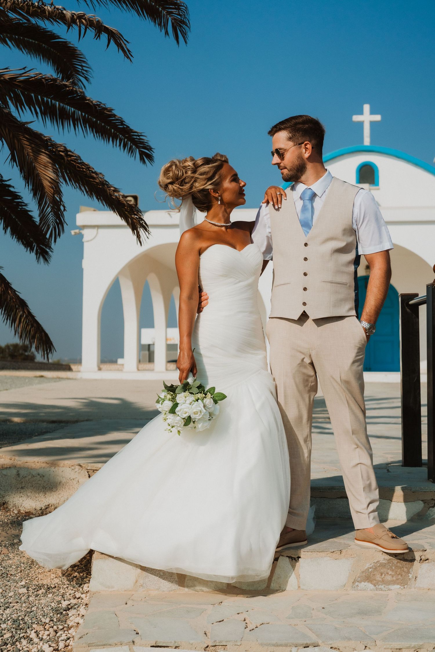 Bride and groom posing in front of a white Greek chapel with blue dome and cross, palm tree, clear blue sky.