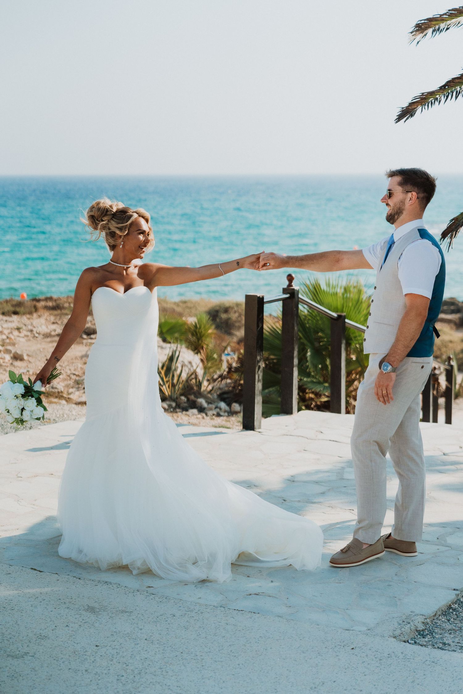 Bride and groom hold hands dancing by the sea, she wears a white mermaid gown, he wears a blue vest and tan pants.
