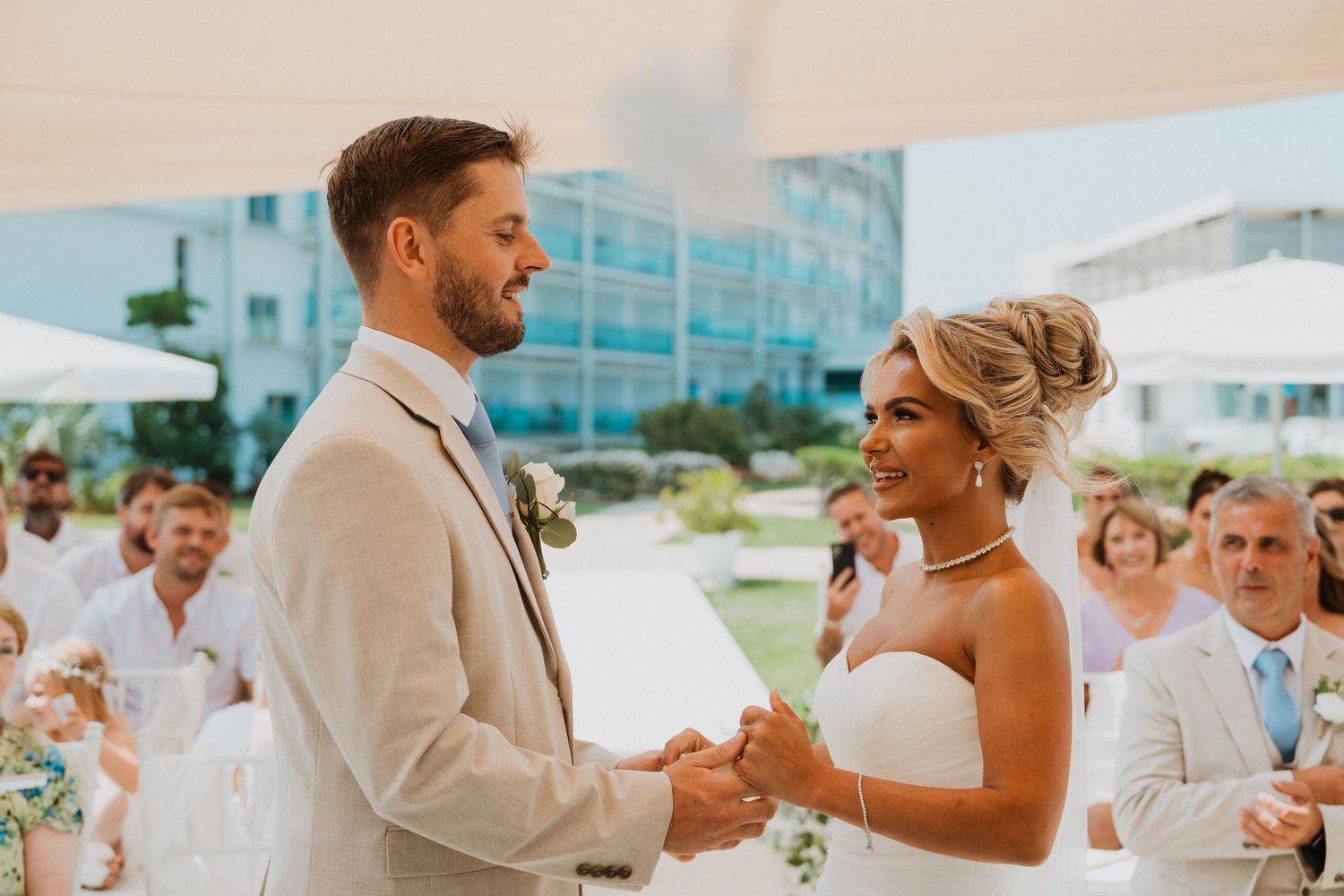 Couple exchanging vows at outdoor wedding ceremony, groom in beige suit, bride in white dress, guests seated behind.