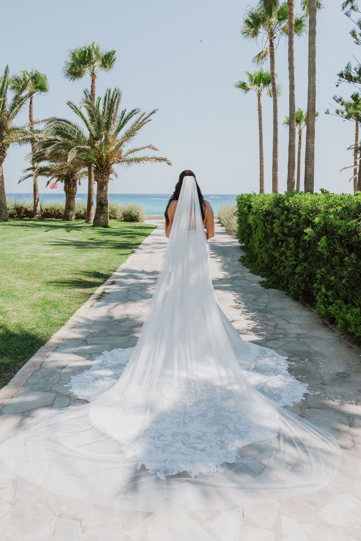 Long cathedral veil trails behind bride on tropical beachside walkway.