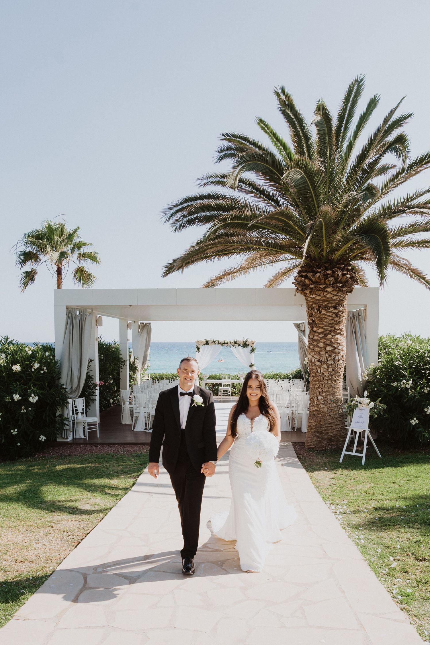 Wedding couple walking along white pathway with palm trees and ocean view.