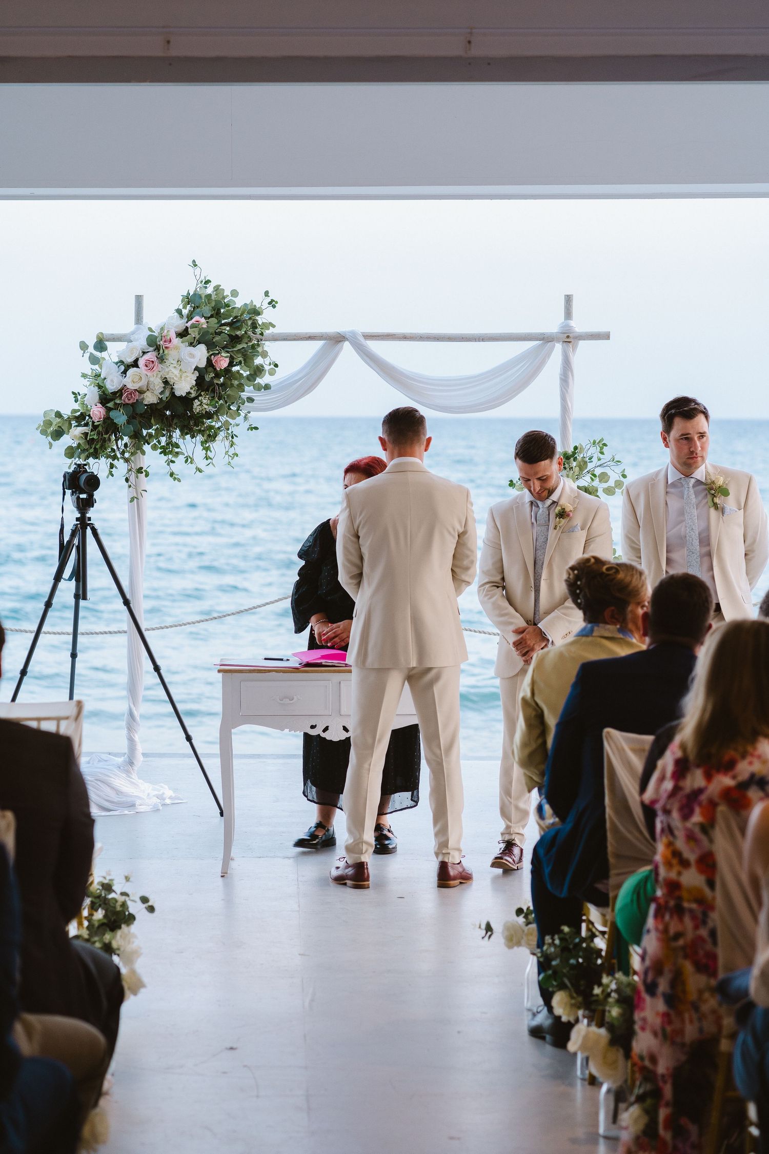 Oceanfront wedding ceremony setup with white arch, floral arrangements, and guests seated on white chairs.