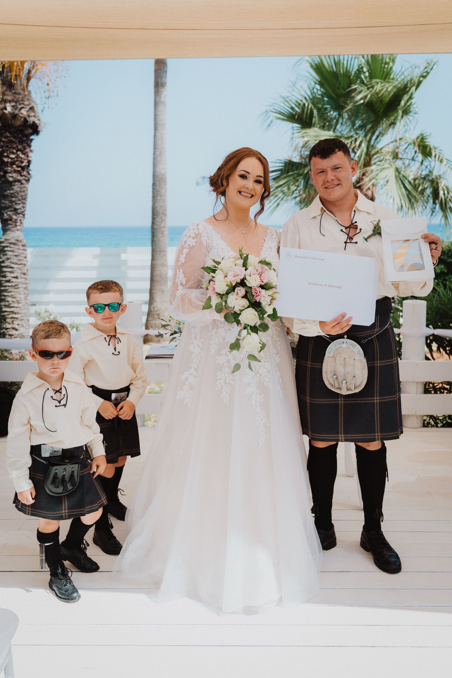 Beach wedding family portrait with Scottish kilts and sunlit palm trees in background.