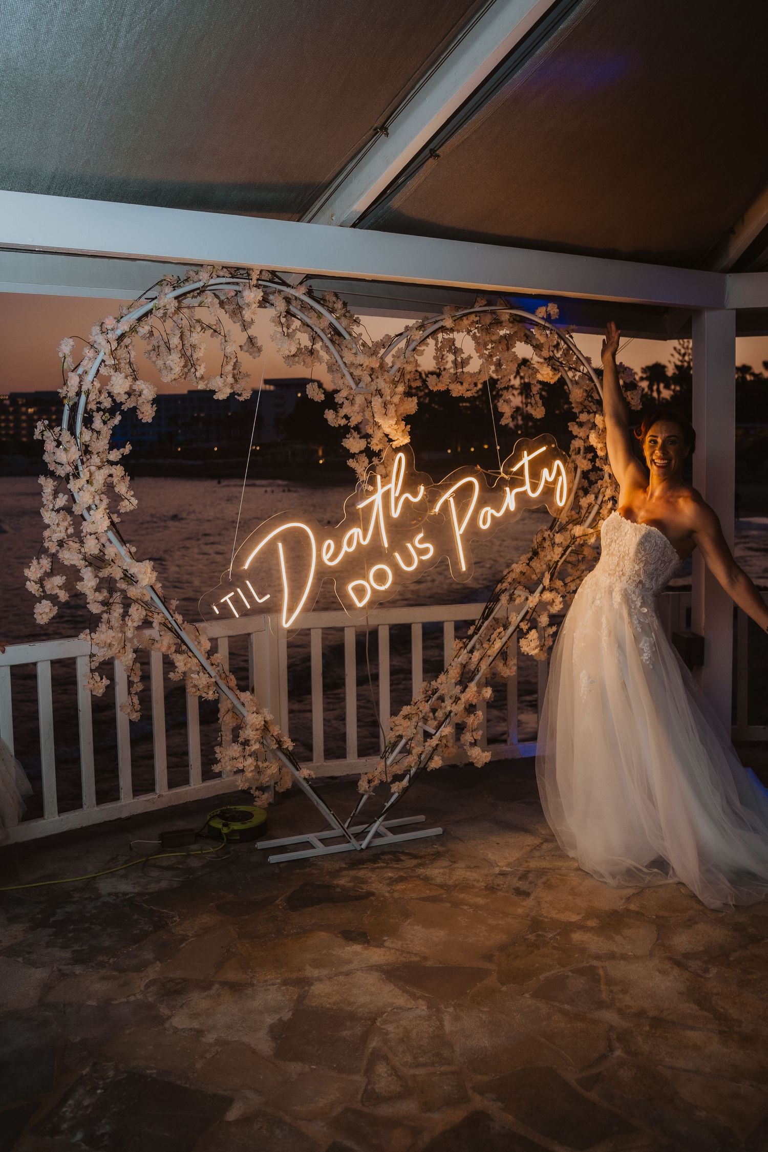 White floral heart arch with neon sign creating romantic ambiance at beach wedding venue.
