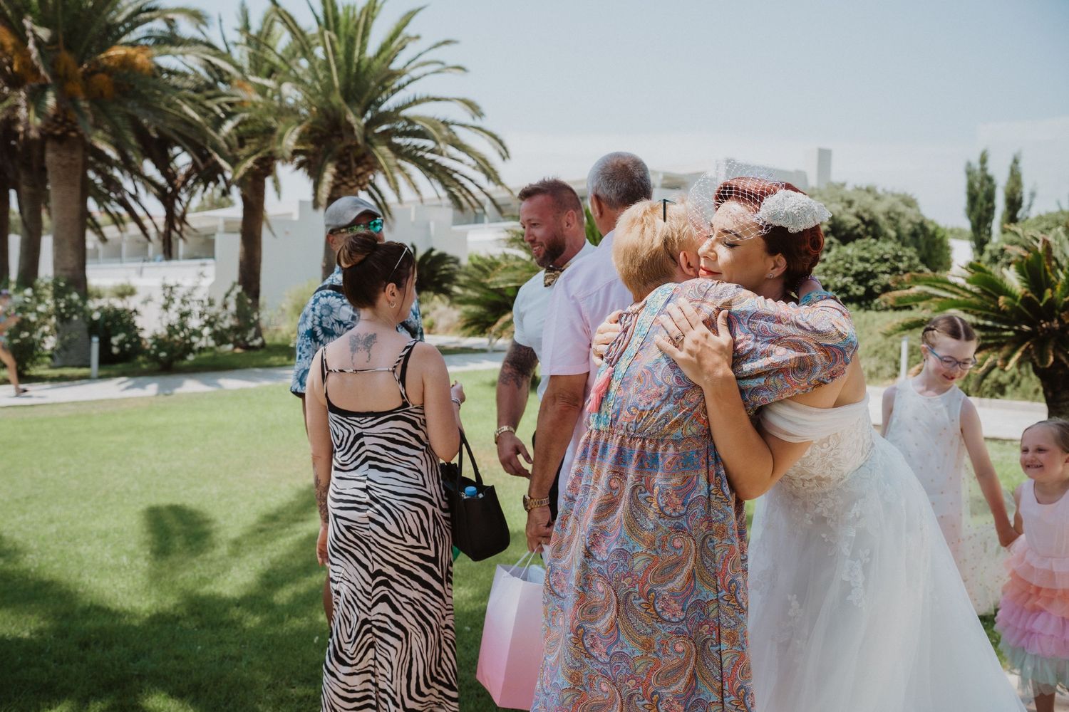 Wedding guests share embraces during outdoor celebration with palm trees in background.