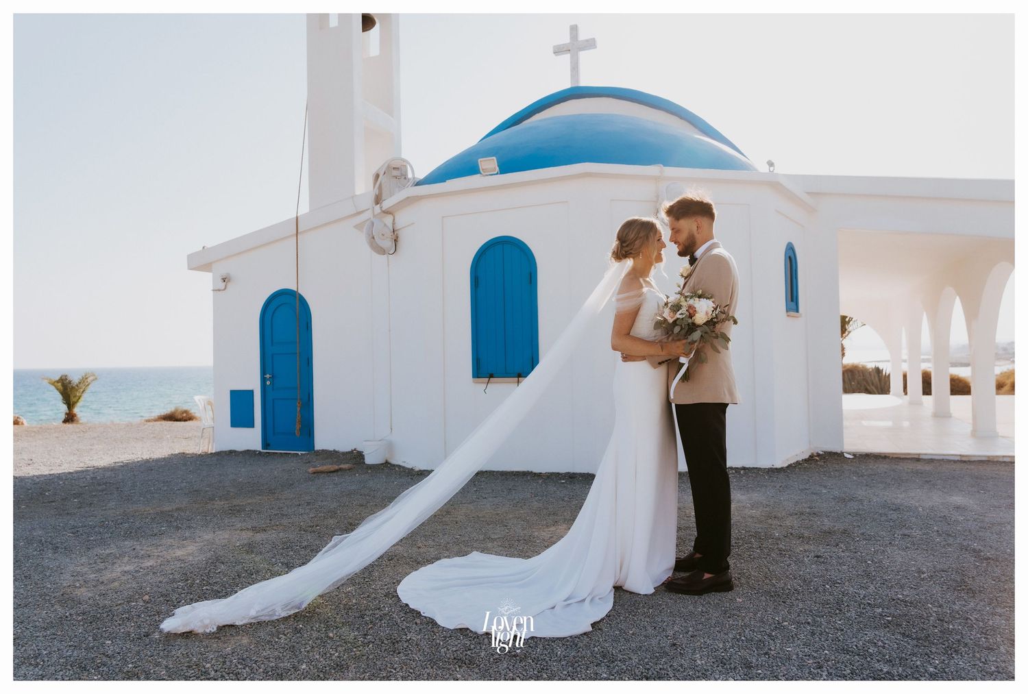Romantic wedding portrait at traditional white Greek church with distinctive blue domed roof.