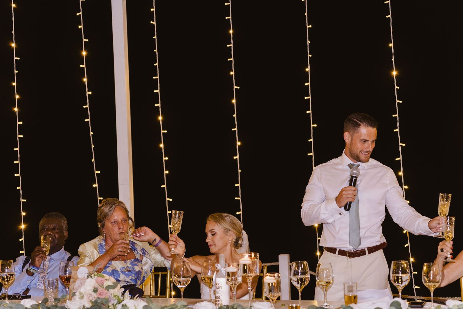 Speaker raises glass for wedding toast against backdrop of twinkle lights at reception.