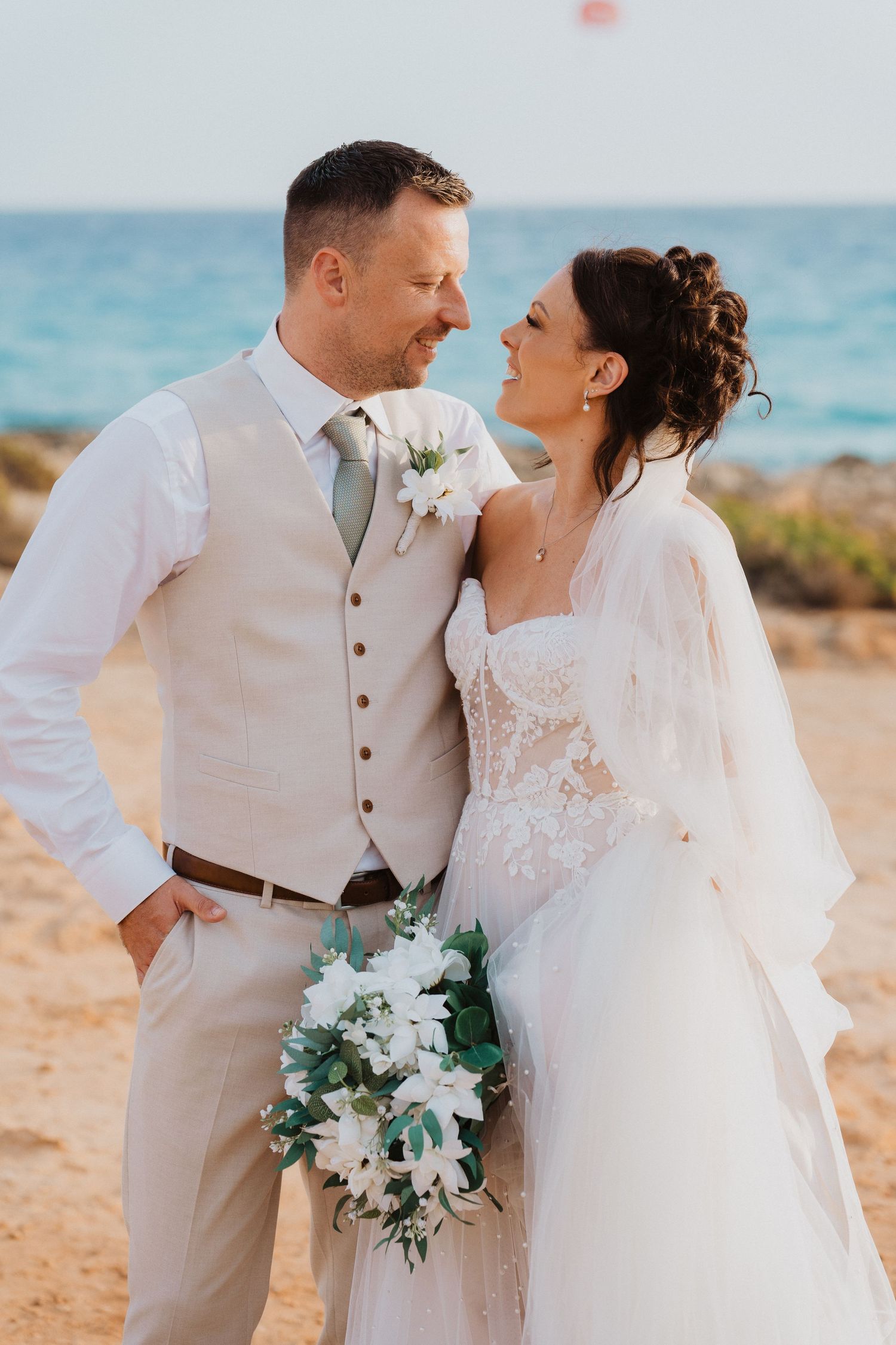 Romantic beach wedding portrait featuring newlyweds in elegant attire with white florals against turquoise ocean backdrop.