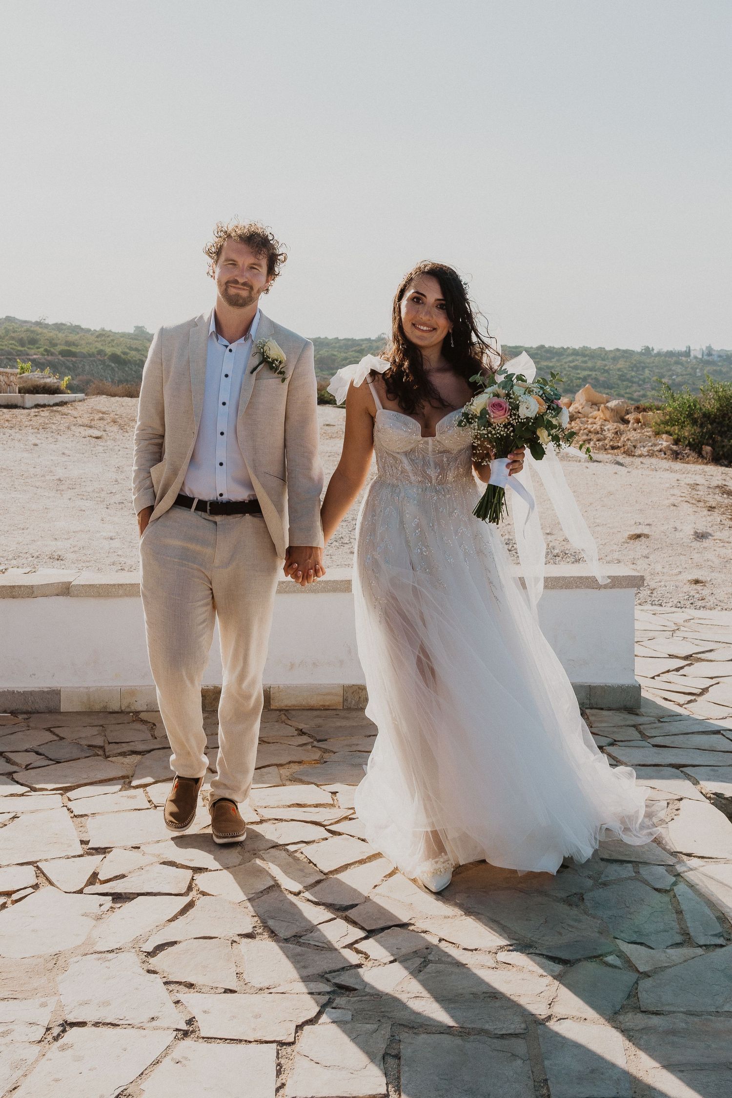 A wedding couple walks hand in hand on a stone patio with a bright sunny backdrop.